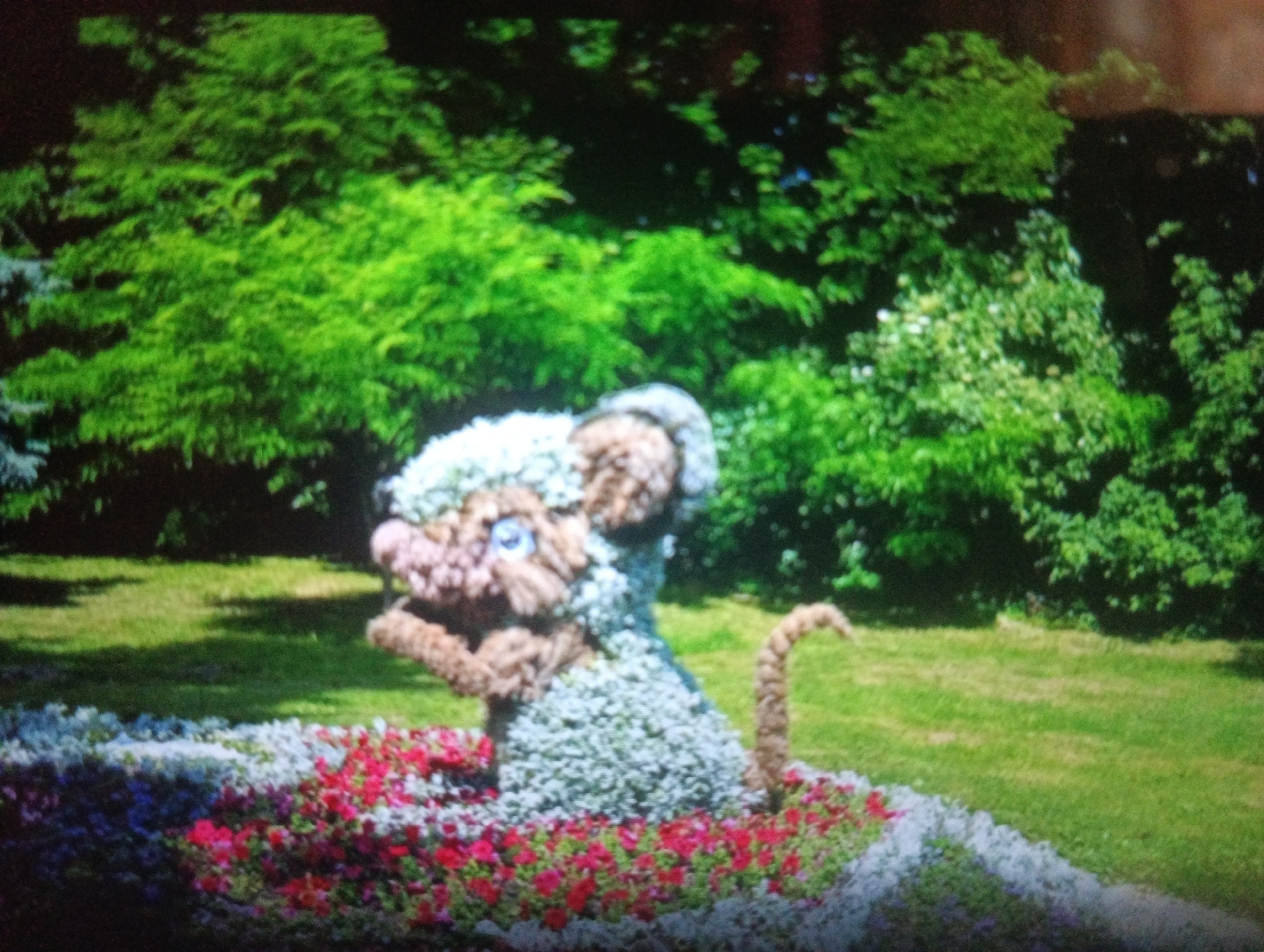 An outdoor scene features a topiary animal, resembling a mouse, positioned in a flowerbed. The topiary is crafted from textured material, with a brown nose, blue eyes, and a fluffy white headpiece. It appears to be seated, with its tail curled upwards. The surrounding flowerbed is densely planted with small red and white flowers. In the background, lush green trees and a grassy lawn are visible, suggesting a park or garden setting. The lighting indicates it is daytime.