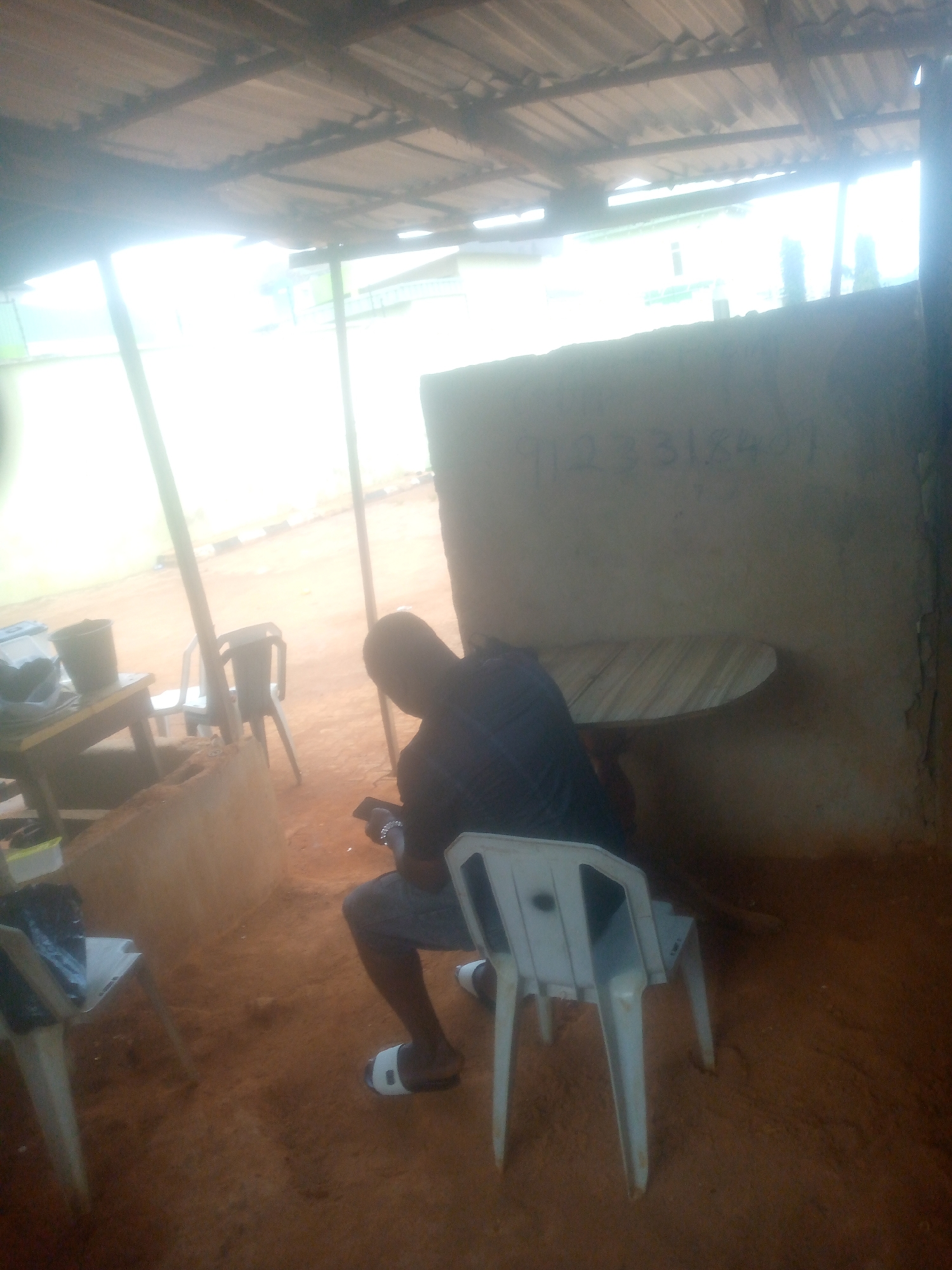 A person is seated in a plastic chair beneath a corrugated metal roof in an outdoor setting. The person is facing away from the camera and is looking at a mobile phone. The ground is red dirt. A wall behind the person has numbers written on it. There are other chairs and a table in the vicinity. Location: Unknown, Unknown.
