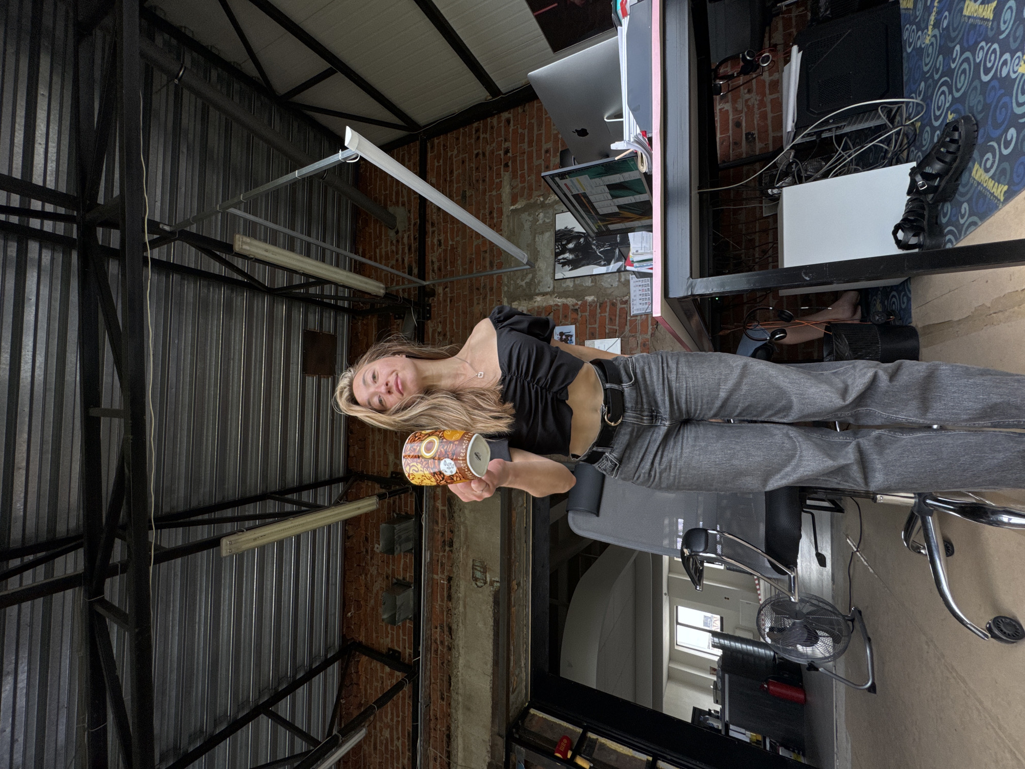 The image depicts a young woman standing in what appears to be an industrial-style loft or studio space. She is holding a decorative mug with a colorful pattern in her right hand, offering it towards the camera. She is wearing a black off-the-shoulder crop top and dark grey high-waisted jeans with a black belt. Her blonde hair is long and wavy.
The environment shows exposed brick walls, a metal truss ceiling with fluorescent lighting, and industrial ductwork. To the right, a desk is set up with a computer monitor, keyboard, and various papers. An office chair is positioned in front of the desk. In the background, an open doorway reveals another room with windows and some storage units. On the floor to the left of the woman, a black oscillating fan is visible. The overall impression is a creative or work-oriented space.
The location is identified as Chaikovsky, Russia.