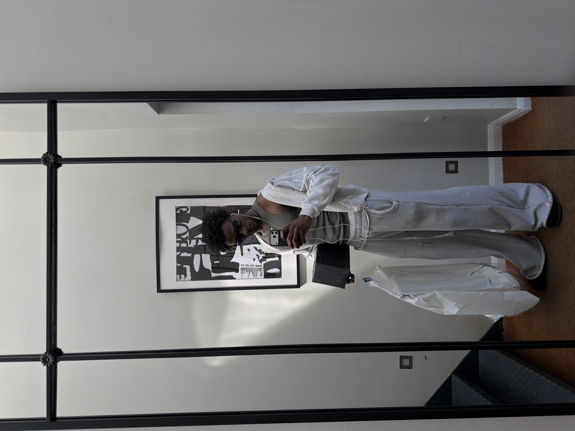 A person is reflected in a full-length mirror with a black metal frame. The person, who appears to be male, is wearing a light grey sweatsuit with a matching hoodie, a grey tank top, black shoes, and sunglasses. He is holding a smartphone and appears to be taking a selfie. He has earphones in.
Behind the person in the reflection, there is a black and white framed art print on the wall. Next to the person is a black box and a shirt in plastic wrap. There is a staircase visible in the bottom right corner of the reflection. The flooring appears to be wooden.
The location is not discernible from the image.