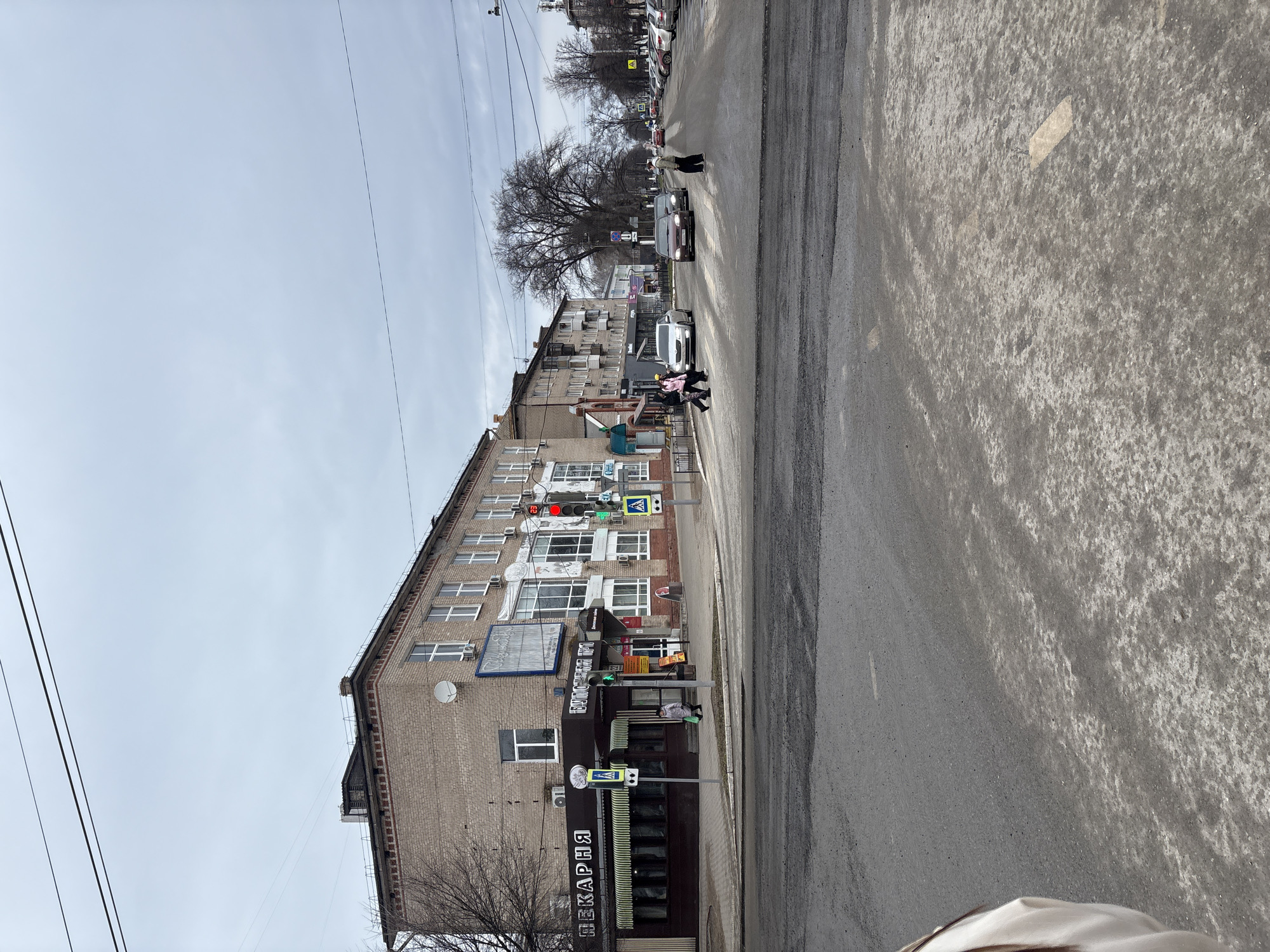 The image shows a street scene in Chaikovsky, Russia. The view is from the sidewalk looking across the street towards a brick building. The building appears to be multi-story with shops at street level, including a bakery ("ПЕКАРНЯ"). The road surface is asphalt and shows signs of wear. There are pedestrian crosswalks visible. There are also a couple of pedestrians at the crosswalk. In the street, one car is visible, other cars and pedestrians are further down the street. The sky is overcast. Power lines run along the street. Trees are present, some are bare.