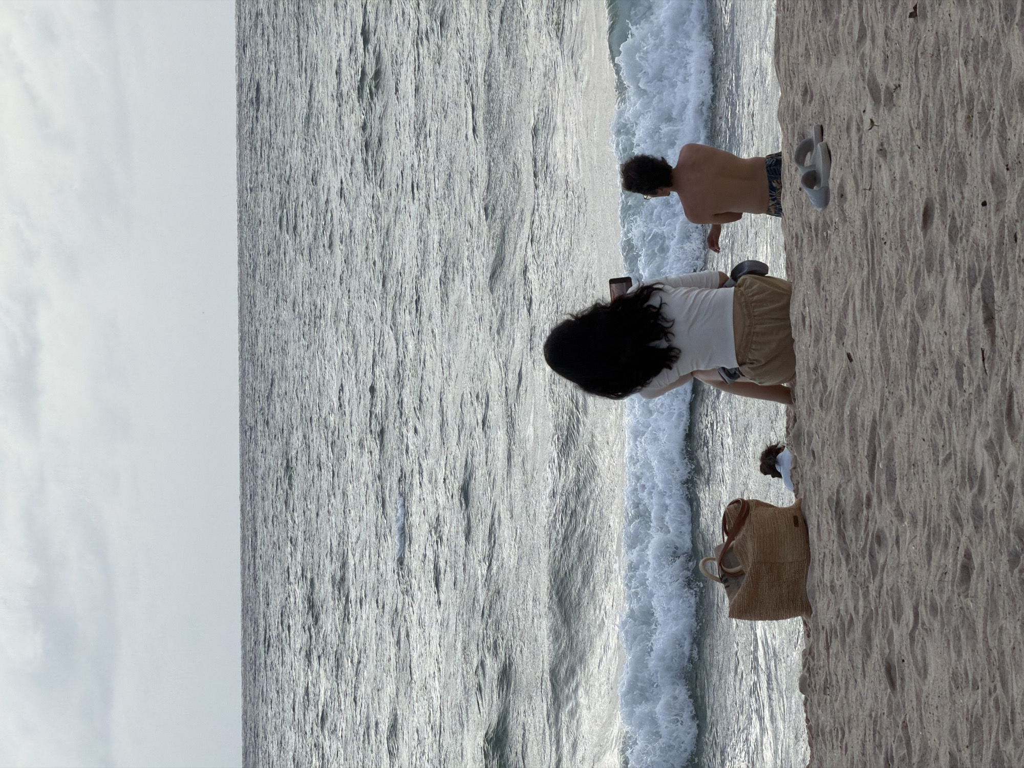 The image depicts a scene at a beach in Choeng Thale, Thailand. A man and a woman are standing near the shoreline. The woman is holding a mobile phone. A pair of grey sandals are on the beach. A woven bag is on the sand near the woman. Ocean waves are breaking. The sky appears overcast.