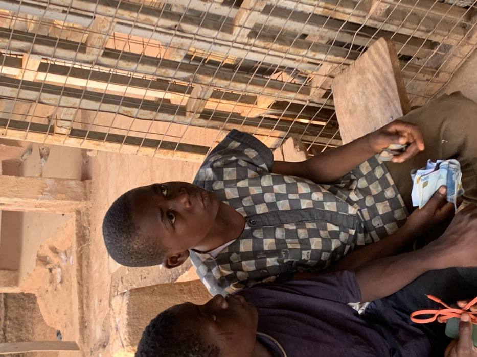 This image depicts two young males in an outdoor setting in Ube, Nigeria. The primary subject, a male estimated to be 8-12 years old, possesses short, dark hair and wears a short-sleeved collared shirt with a checkerboard pattern of light and dark grey squares. He is seated on a light-colored wooden surface. In his left hand, he holds a stack of various multi-colored banknotes. His gaze is directed towards the right side of the frame.
Positioned to his left and partially visible is a second male, also appearing to be 8-12 years old, wearing a dark blue short-sleeved top. This individual exhibits distinct linear scarification marks on his left cheek. He holds an orange braided cable in his left hand and a dark rectangular device in his right hand.
The background consists of exposed reddish-brown sandy earth and several weathered building structures, suggestive of aged concrete or mud-brick construction. To the right of the seated individuals, a tall, rectangular metal grid structure, possibly a gate or security screen, stands vertically, casting sharp linear shadows across the scene. The entire scene is illuminated by strong natural sunlight, creating a warm hue.