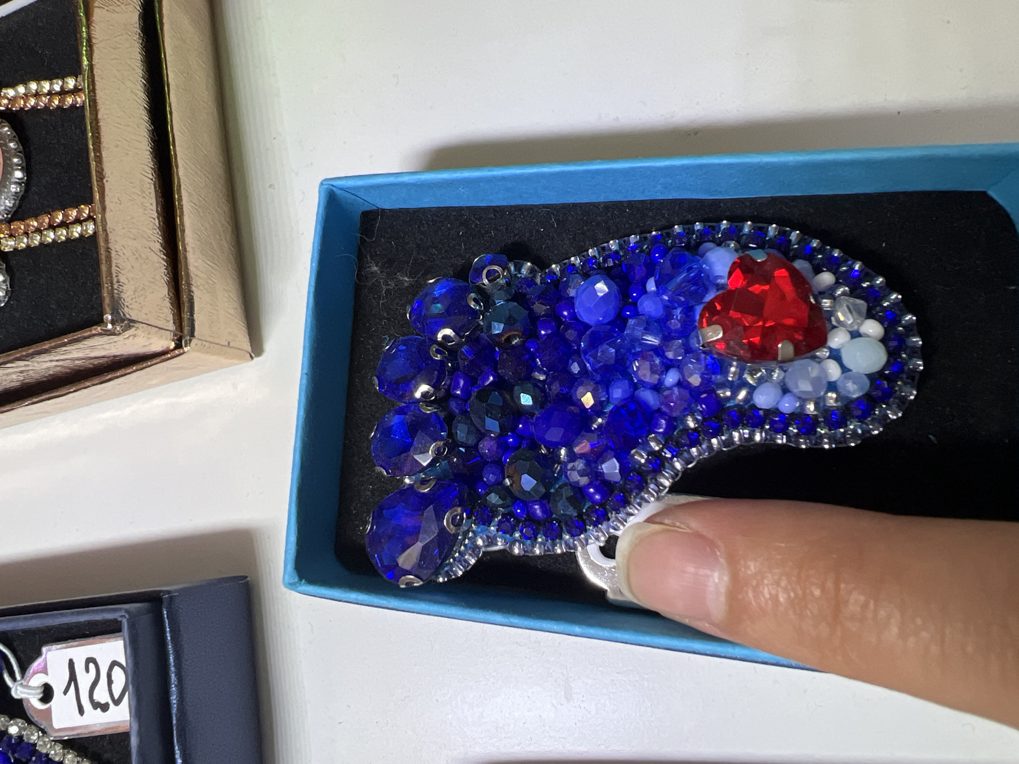 A close-up shot depicts a handcrafted brooch nestled in a small, bright blue box with a black interior. The brooch is composed primarily of dark blue beads and crystals, clustered together in a somewhat irregular shape reminiscent of a paw print. A central red, heart-shaped stone is surrounded by smaller white beads. The brooch appears to be secured with a silver clasp. A portion of a human finger is visible, holding the brooch. To the left of the box, a gold-colored box is partially visible, showcasing jewelry with metallic accents and rhinestones. In the lower-left corner, a navy-blue box contains a price tag marked "120." The background is plain white. There is no evidence that the image was taken in Chaikovsky, Russia.
