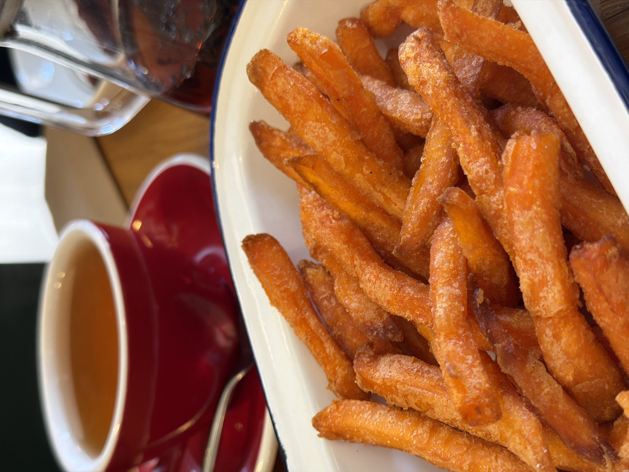 A close-up, high-angle shot captures a plate of sweet potato fries next to a red cup of tea. The fries are piled in a white dish with a blue rim. They appear golden brown and possibly sugared or lightly salted, with a slightly rough texture suggesting they are either baked or fried. The tea cup is red with a white interior, and it is filled with a light-colored liquid. Part of a glass serving container and a surface, possibly a wooden table, are visible in the background. The scene appears indoors. No people are visible. The location is unknown.