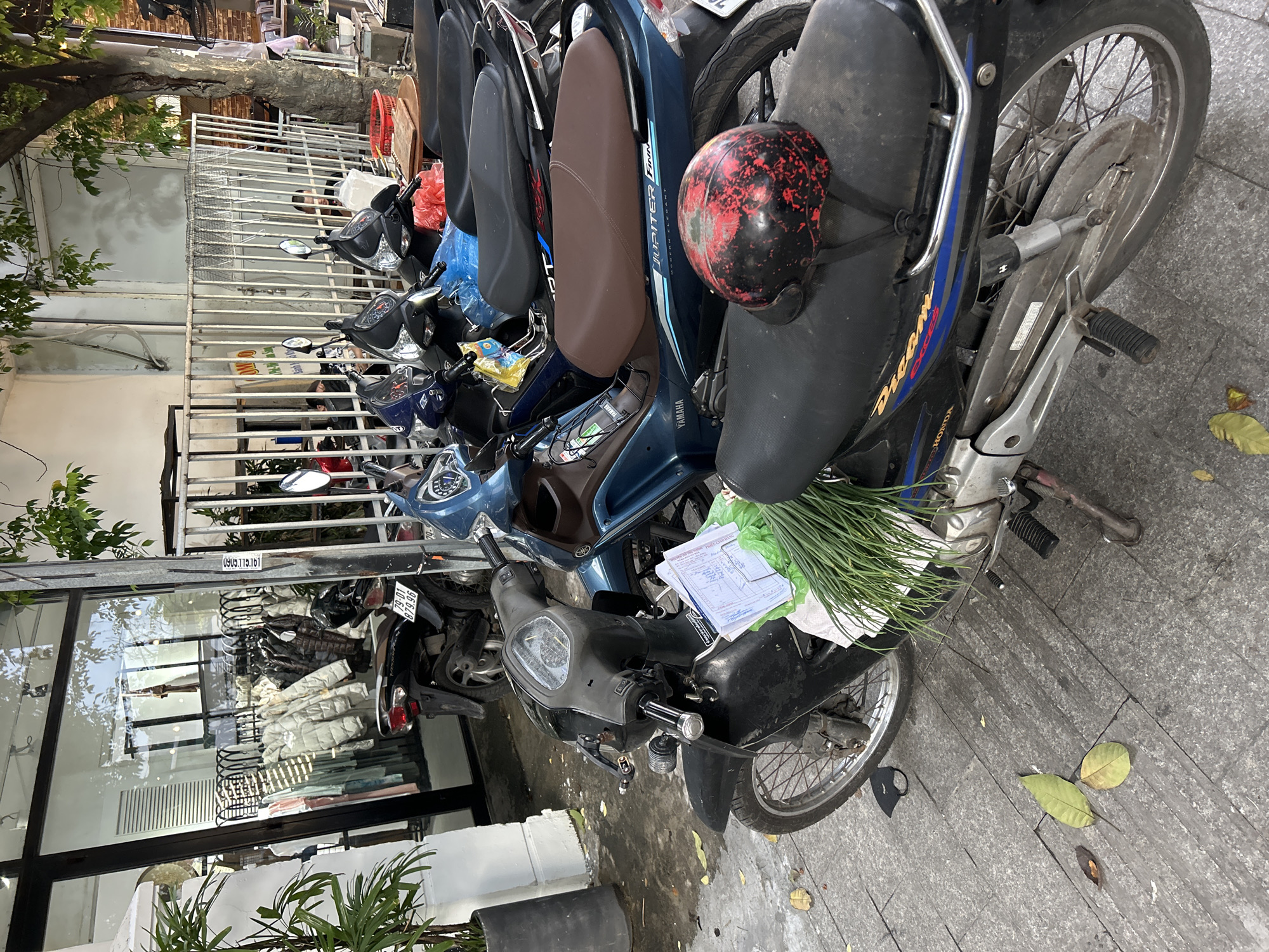 The photograph captures a daytime street scene in Nha Trang Ward, Vietnam, bustling with parked motorbikes along a paved sidewalk. The bright, even lighting suggests it is either morning or afternoon.
A prominent blue Yamaha Jupiter Finn scooter with a brown seat is visible among the many parked vehicles. On the seat of a black motorbike in the foreground, a bundle of fresh green onions or chives is placed next to some papers, possibly market receipts or delivery documents. A black helmet, adorned with red paint splatters, rests on the seat of an adjacent motorbike. Further down the row, a black Honda Dream Exces underbone motorbike is identifiable by its branding. A black face mask lies on the grey sidewalk tiles, alongside a few fallen green leaves.
To the left, a modern building features a large glass storefront, revealing clothes on hangers and rolled fabric inside, suggesting a retail shop. Above this window, a metal beam displays a Vietnamese phone number, "0903.119.161". A white metal gate separates the sidewalk from the property, behind which glimpses of individuals are visible. A motorbike license plate, "79-D1 079.96", is clearly seen, with "79" indicating Khánh Hòa province, where Nha Trang is located. A lush tree with green foliage partially shades the left side of the frame. The scene portrays a typical urban environment in Vietnam, characterized by dense motorbike parking and active street-level commerce.