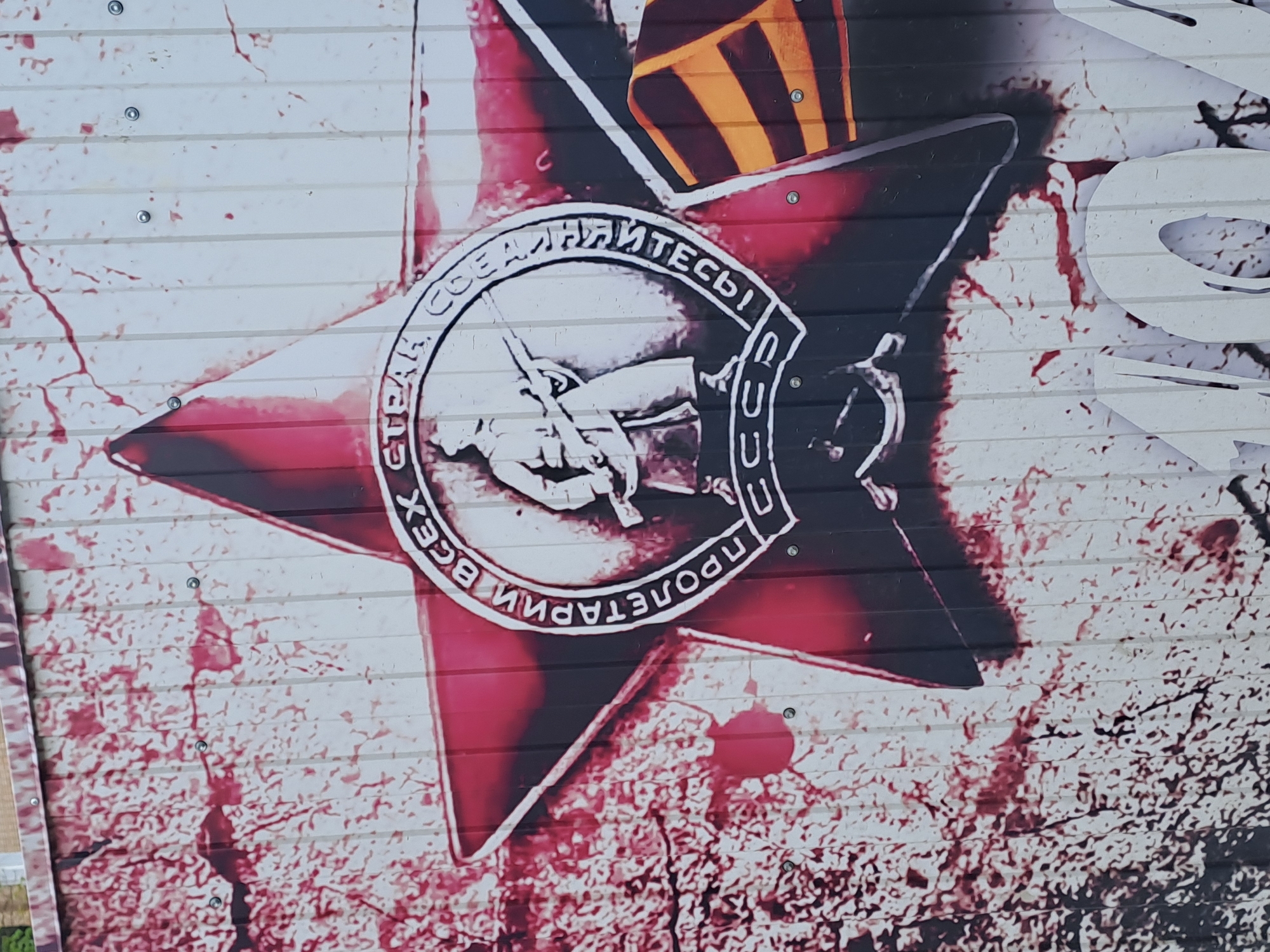The image shows a close-up of a painted wall with a stylized Soviet-era propaganda motif. The central element is a red star with a white border. Inside the star is a circular emblem featuring two hands holding tools (likely a hammer and sickle, though details are obscured). The circle is inscribed with text in Russian. Above the emblem is what appears to be a black and orange striped ribbon. The paint on the wall is cracked and textured. The background is a speckled pattern. Based on the context, the image is likely located in the city of Elektrostal, Russia.