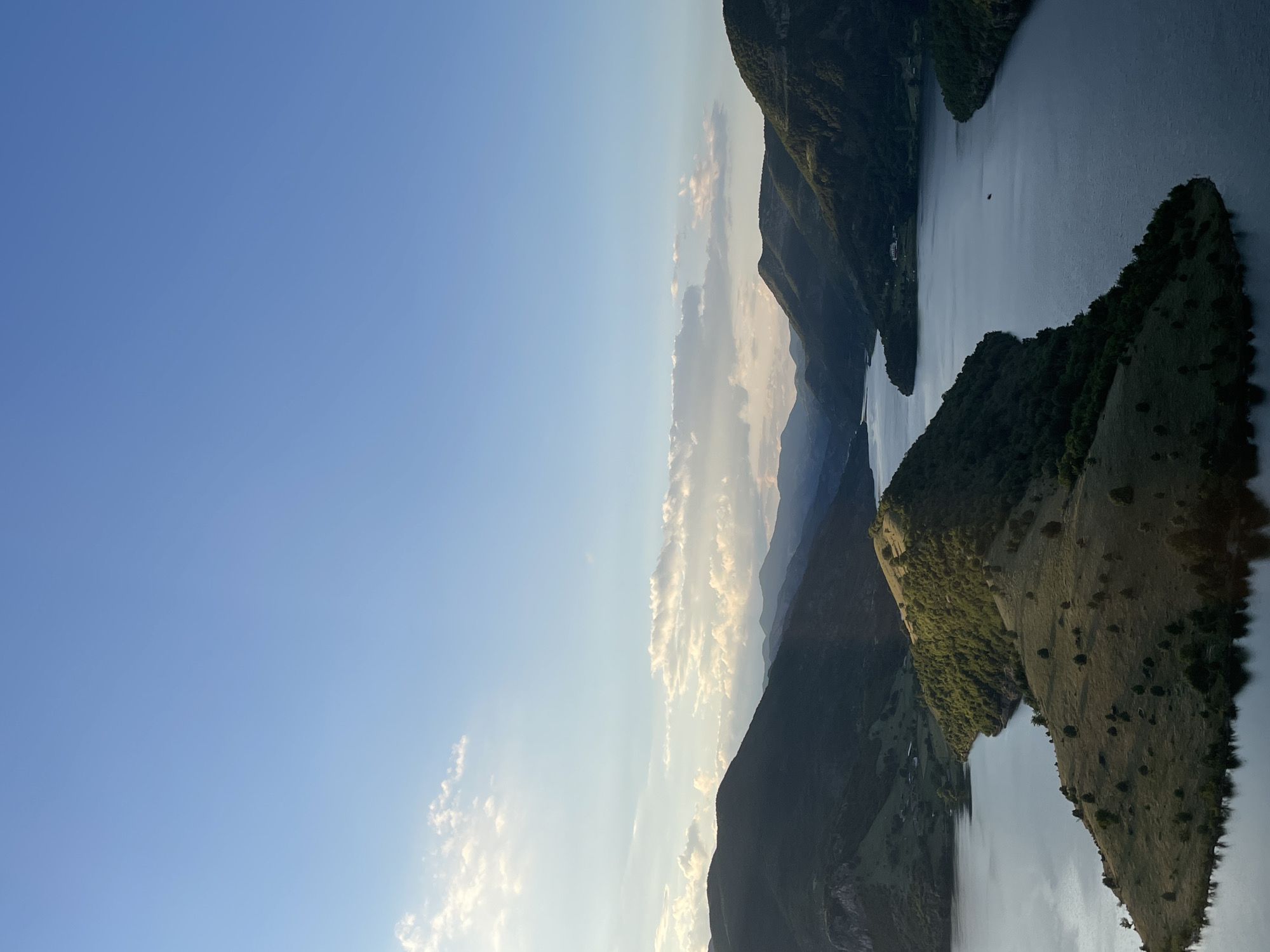 This image captures an expansive view of a serene lake nestled amidst rolling, verdant hills. The sky above is a clear, bright blue, with a scattering of soft, white clouds, particularly on the left side where the sun's rays create a gentle, luminous glow. The terrain is characterized by steep, forested slopes that rise from the water's edge, with some lighter, grassy areas visible on the more prominent peninsulas. A distinct, sharply defined peninsula juts into the lake in the foreground, its grassy surface dotted with scattered trees. The water itself is a deep, placid blue, reflecting the surrounding landscape and sky, with a subtle ripple on its surface. In the mid-ground, a small, dark red object, possibly a boat or a buoy, is visible on the water. In the distance, a series of layered hills fade into a soft, hazy silhouette against the horizon. The overall impression is one of natural tranquility and a vast, unspoiled landscape, suggesting a remote or sparsely populated area. The image is taken from a high vantage point, offering a panoramic perspective of the geographical features. The location is identified as Borovitsa, Bulgaria.