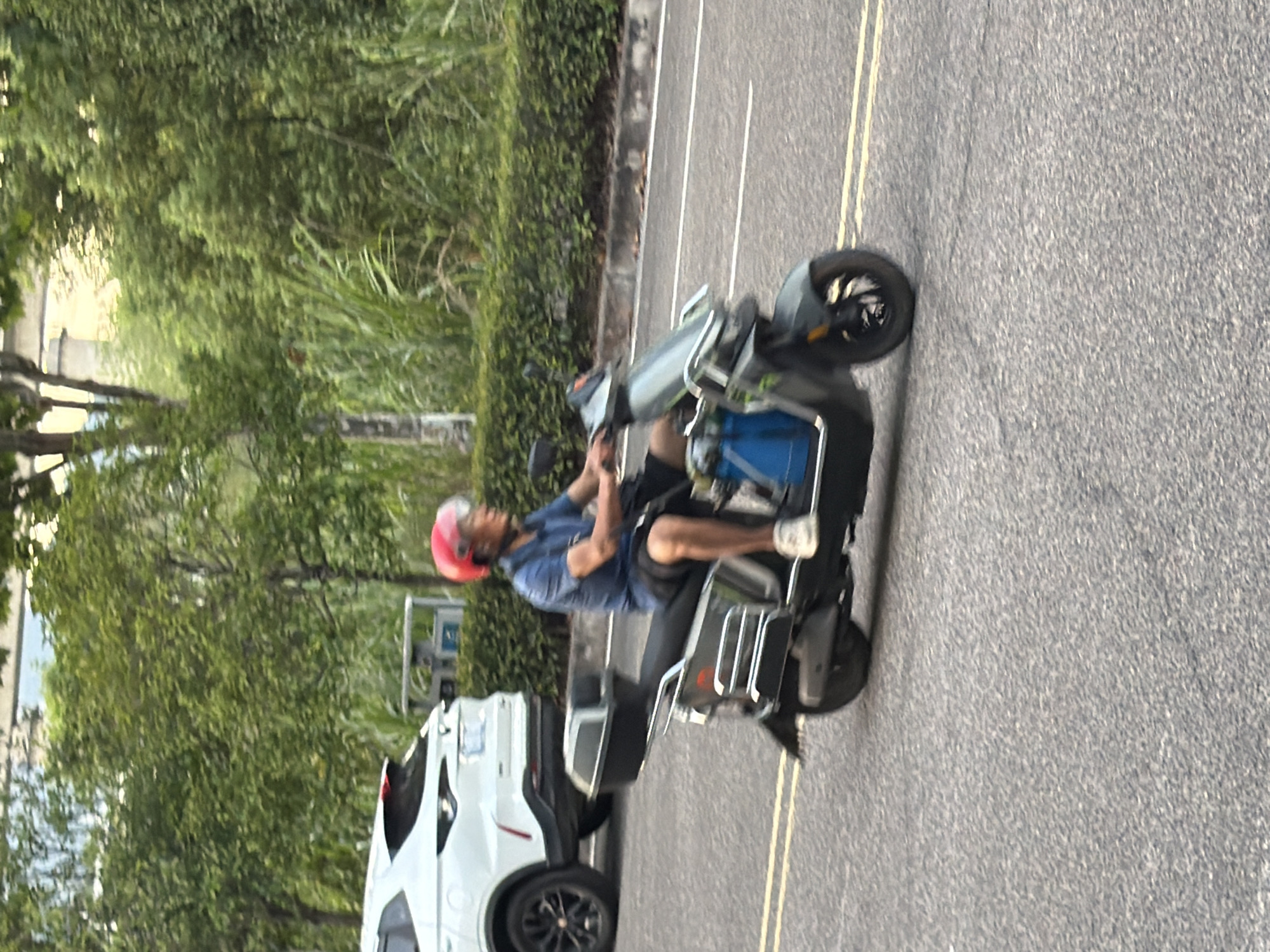 An outdoor, eye-level, medium shot captures a man in a blue shirt and black shorts riding a scooter on a street in Jiangjin, China. He is wearing a red helmet and white shoes. The scooter is dark gray with a black storage box. A white SUV is parked on the left side of the road, behind the scooter. The background consists of green trees and shrubs lining the street. The asphalt road has yellow double lines marking the lanes. The image is slightly blurred, suggesting motion.