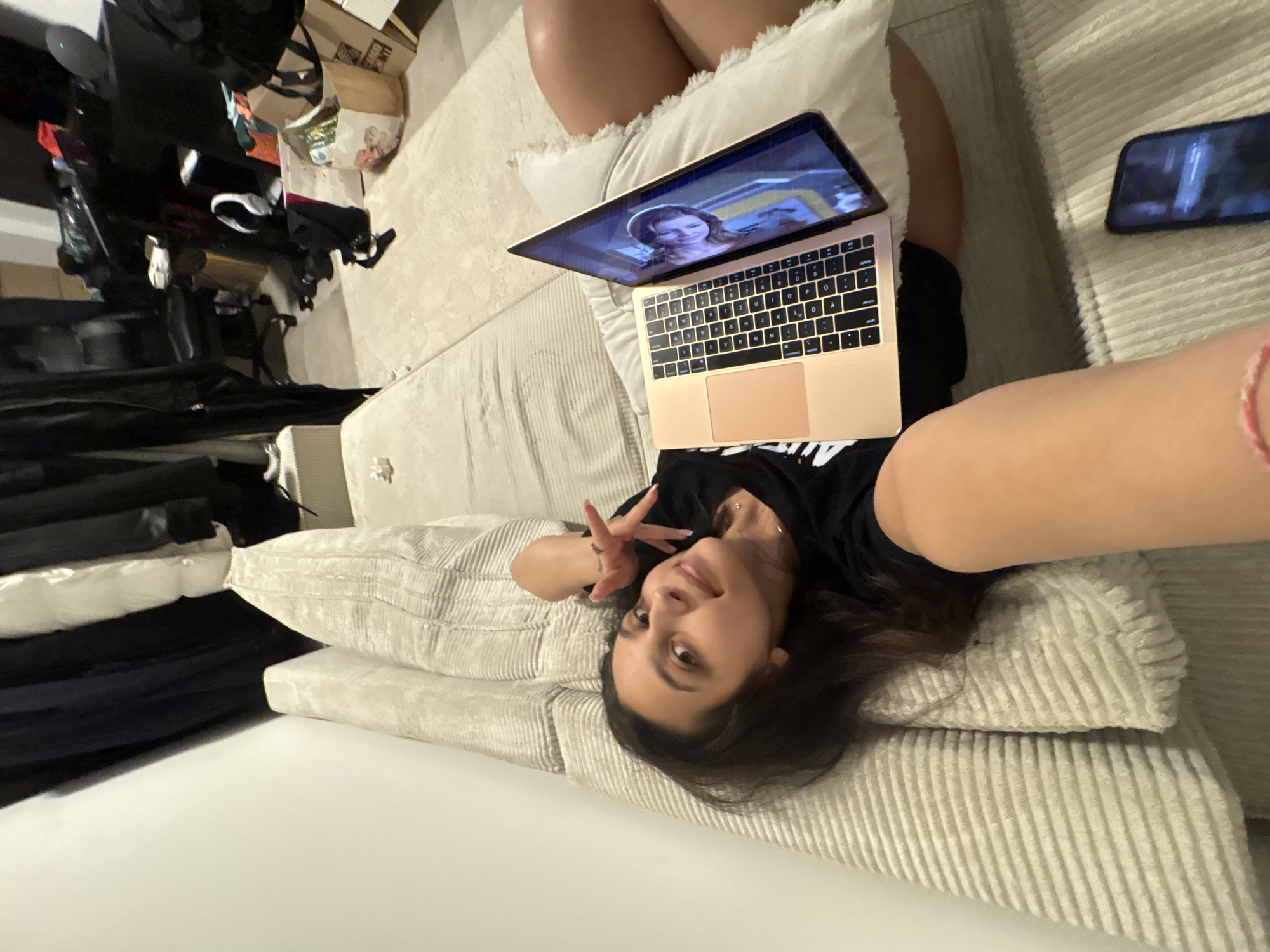 The image depicts a young woman lying on a beige sofa, using a laptop. She is wearing a black t-shirt and appears to be indoors, possibly in an apartment or dorm room. The background shows a cluttered space with clothing racks, furniture, and bags, suggesting a living area. There is also a smartphone on the sofa next to her. The overall lighting is dim. The location within the city of Mönchengladbach, Germany is not discernible from the image.