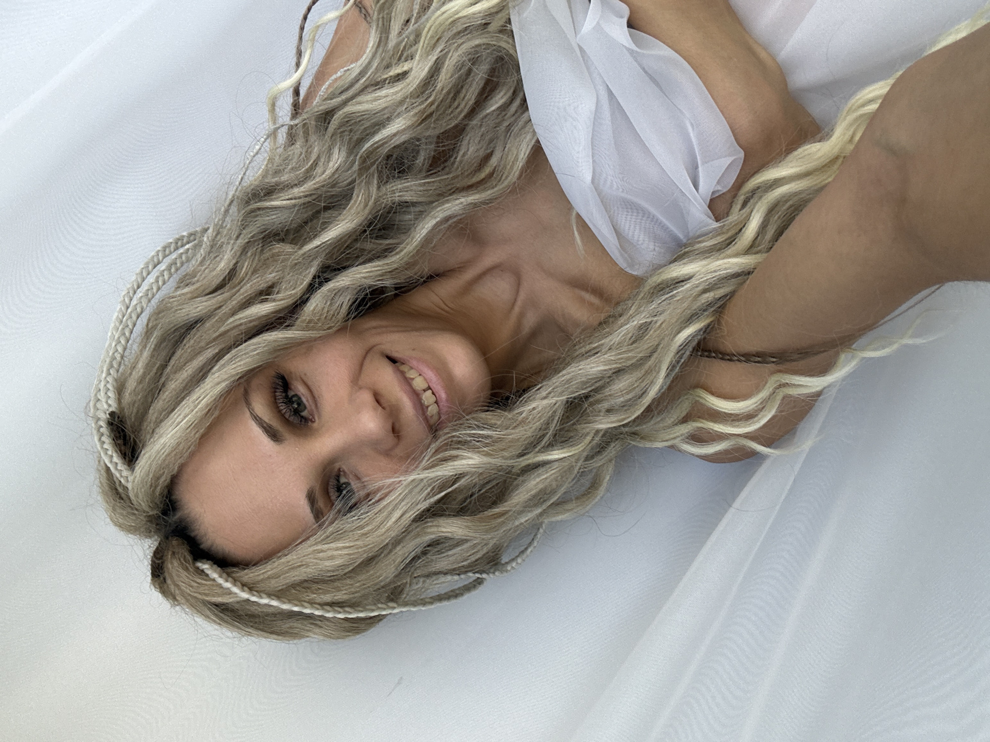 A woman with blonde, braided hair lies on a white, sheer fabric surface. She is smiling slightly and looking at the camera. A piece of white fabric is draped across her upper body. The location is not specified as Rusanovo, Russia; there is no information in the image to suggest this.