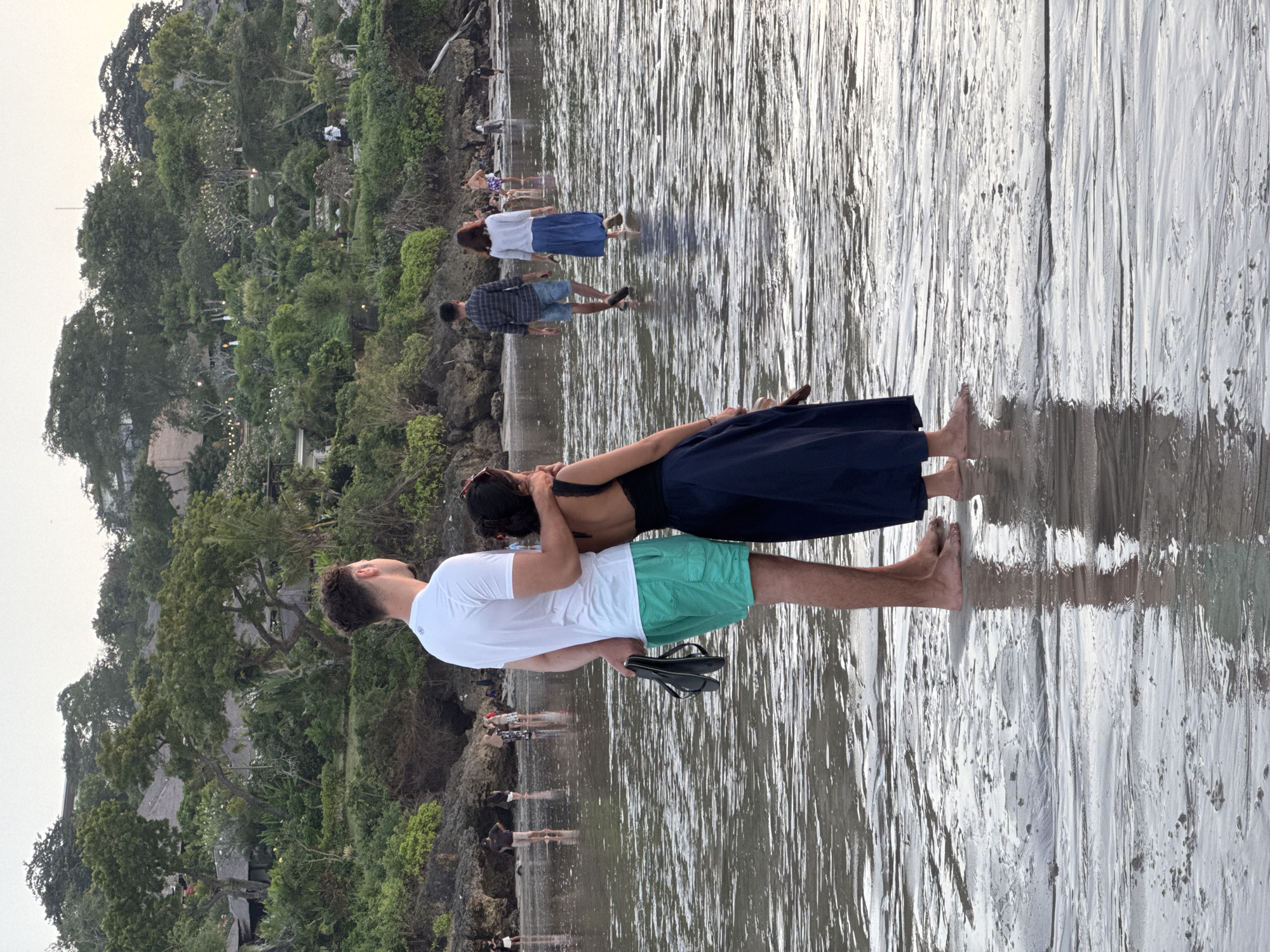 A man and woman stand in shallow water on a beach, facing away from the camera. The man is wearing a white t-shirt and green shorts, and he is holding a pair of black flip-flops in his left hand. The woman is wearing a dark blue dress. The man has his right arm around the woman's shoulders. In the background, other people are walking on the beach and in the water. The beach is backed by a lush, green hillside with buildings and trees. The sky is overcast. The scene appears to be sunset or early evening. The location is likely Kuta Selatan, Bali, Indonesia.