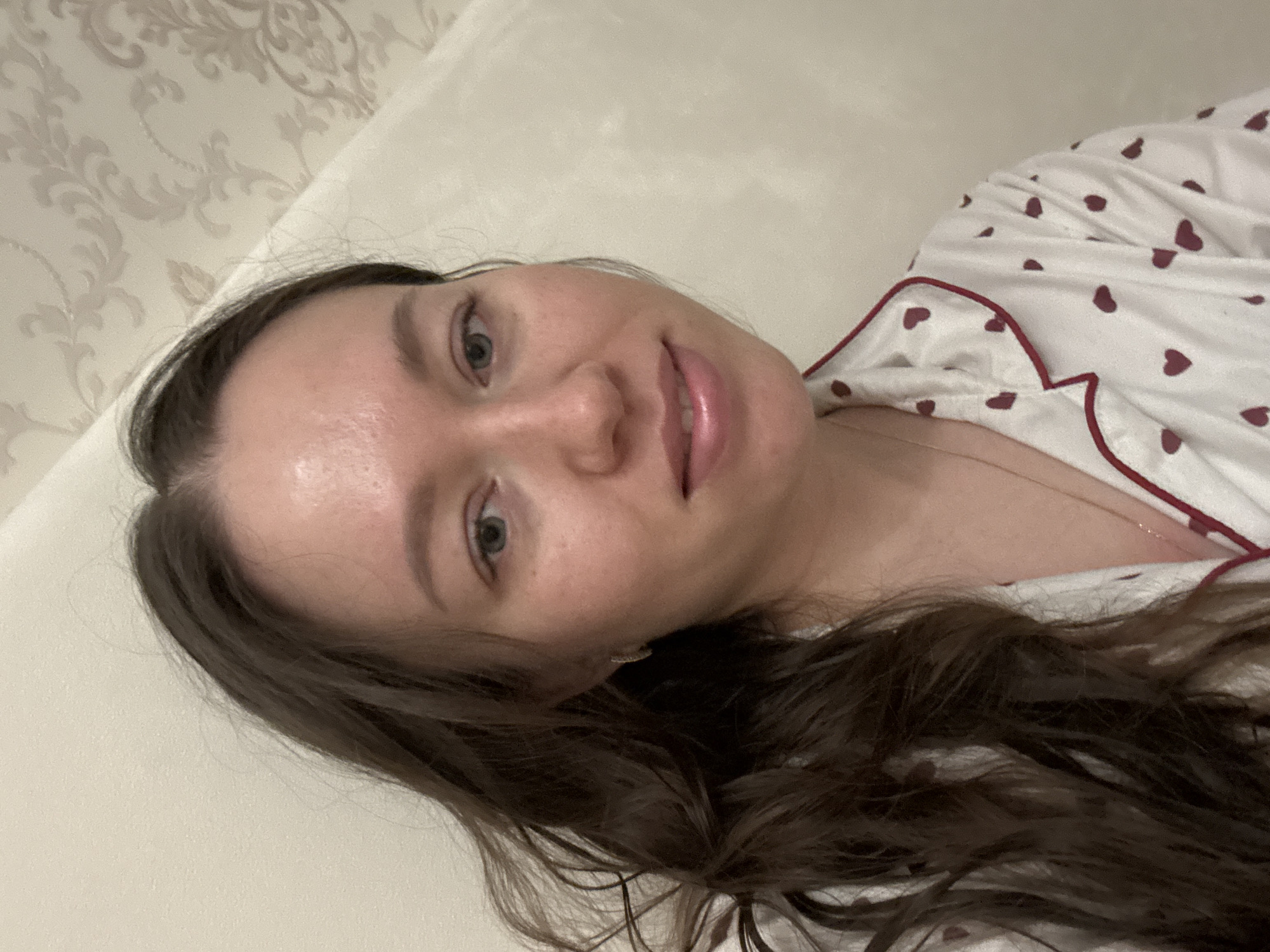 A young woman is lying on a bed in a room with light-colored walls. The headboard of the bed is a light beige color with a decorative pattern. The woman has long, wavy brown hair and fair skin. She is wearing a white pajama top with small red heart patterns and red piping. She is facing the camera with a slight smile. The location appears to be indoors, potentially a residential setting. It is not possible to confirm the location as Ferma, Russia based on the image alone.