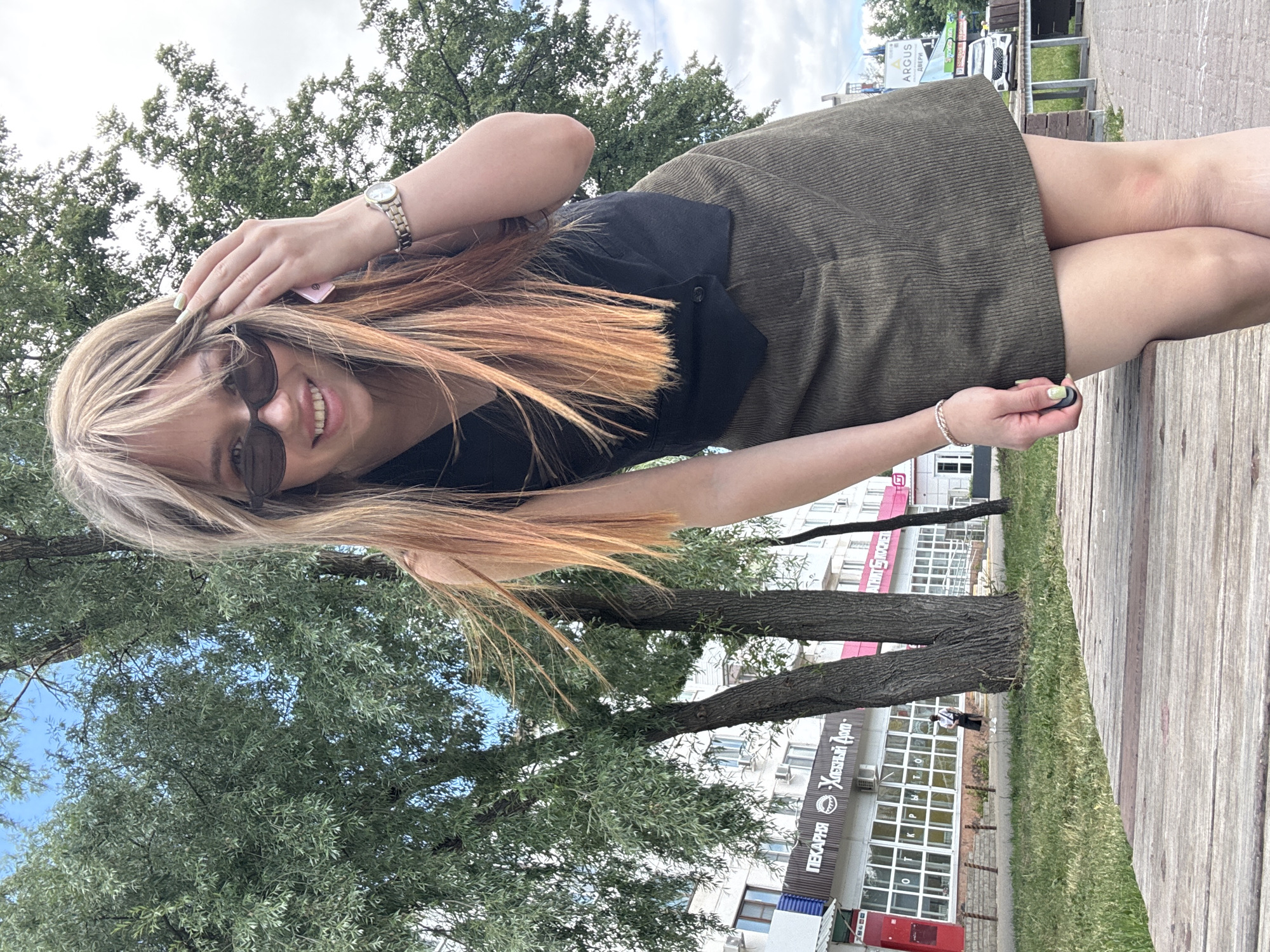 A young woman with blonde hair and sunglasses is standing outdoors in front of trees and buildings, likely in Chaikovsky, Russia, based on the Cyrillic text on the building. She is wearing a black vest, a green corduroy skirt, and a watch. She is smiling and touching her hair. The foreground is a wooden surface. A street and some cars are visible in the background.