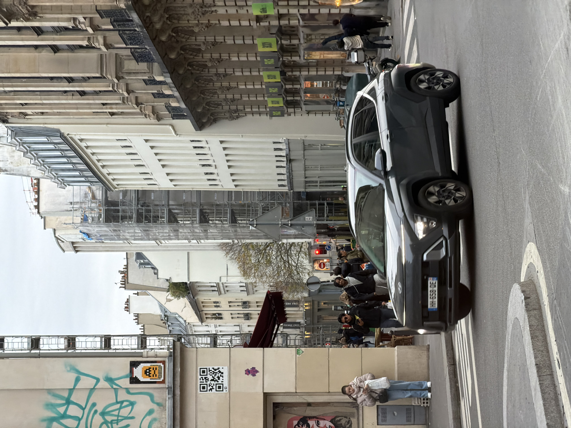 This image depicts a bustling urban street scene in Paris, France, under an overcast sky.
In the foreground, a dark grey Hyundai Kona SUV, bearing the French license plate HH-658-DE, is centrally positioned on the asphalt road, executing a right turn with its headlights illuminated. A curved granite curb delineates the edge of the street on the left.
The left sidewalk features a light-colored building wall adorned with green spray paint graffiti at the top. Below the graffiti, a pixel art mosaic depicting a yellow potion bottle with a skull face and the word "INVADER" is affixed. Further down, a prominent black and white QR code sticker is visible, followed by a small purple pixel art object and an even smaller green diamond sticker. A large poster with a stylized red and white face is partially visible behind the woman on the sidewalk. A young woman, dressed in a light-colored padded jacket and jeans, stands on this sidewalk, looking down at her smartphone while holding a white bag. A grey utility box is situated beside her. Further back on the left, a red awning extends over a storefront, with outdoor cafe seating visible beyond.
In the middle ground, multiple pedestrians are observed walking on both sidewalks and crossing the street. A man in a dark jacket and sunglasses is walking on the left sidewalk, also engrossed in his phone. A red traffic signal is visible on the right side of the street, indicating a stop.
The right side of the street is lined with multi-story buildings, including one with an ornate facade featuring vertical pilasters and small balconies. This building houses several storefronts, each displaying repetitive bright green signs with "BODY" and "FACE" written in white text. A motorcycle is parked on the sidewalk in front of these stores. In the background, numerous buildings, some partially obscured by scaffolding, line the street, culminating in a skyline of varied Parisian rooftops under the grey sky. Trees with minimal foliage are interspersed among the buildings in the mid-distance.
