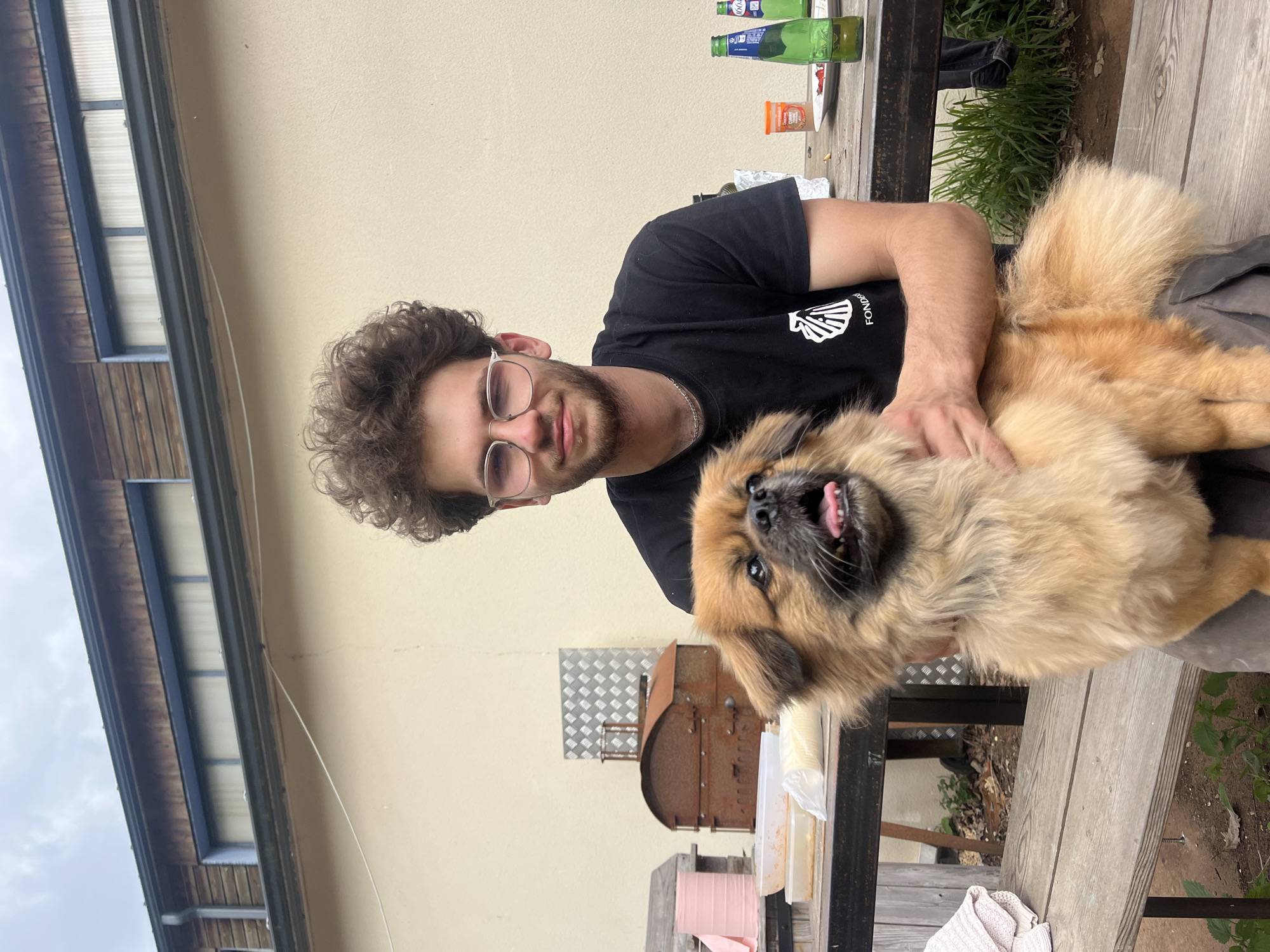 An outdoor scene in Saint-Rémy-lès-Chevreuse, France, depicts a young man holding a fluffy dog. The man, with curly dark hair, a beard, and glasses, is wearing a black t-shirt with a white graphic (partially visible text "FONDE..."). He looks towards the camera with a slight smile, his arm gently around the light brown, long-haired dog. The dog, possibly a Pekingese or similar breed, has darker fur around its snout and looks at the camera with its mouth open, pink tongue visible, conveying a happy and playful expression.
They are positioned next to a rustic wooden table or bench, indicating a backyard or patio setting. On the table, several items are present: two green bottles (one with "100..." on its label, another with a blue label), an orange container labeled "CAY... SAUCE," a white plate with red food residue, and various white plastic items. Below the table, a rusty, barrel-shaped object, likely a barbecue grill or smoker, stands on a metallic checkered sheet. The background shows light-colored building walls and a glimpse of an overcast sky. Patches of grass and dirt are visible near the table's base. The overall mood is casual and cheerful, likely captured during daytime.