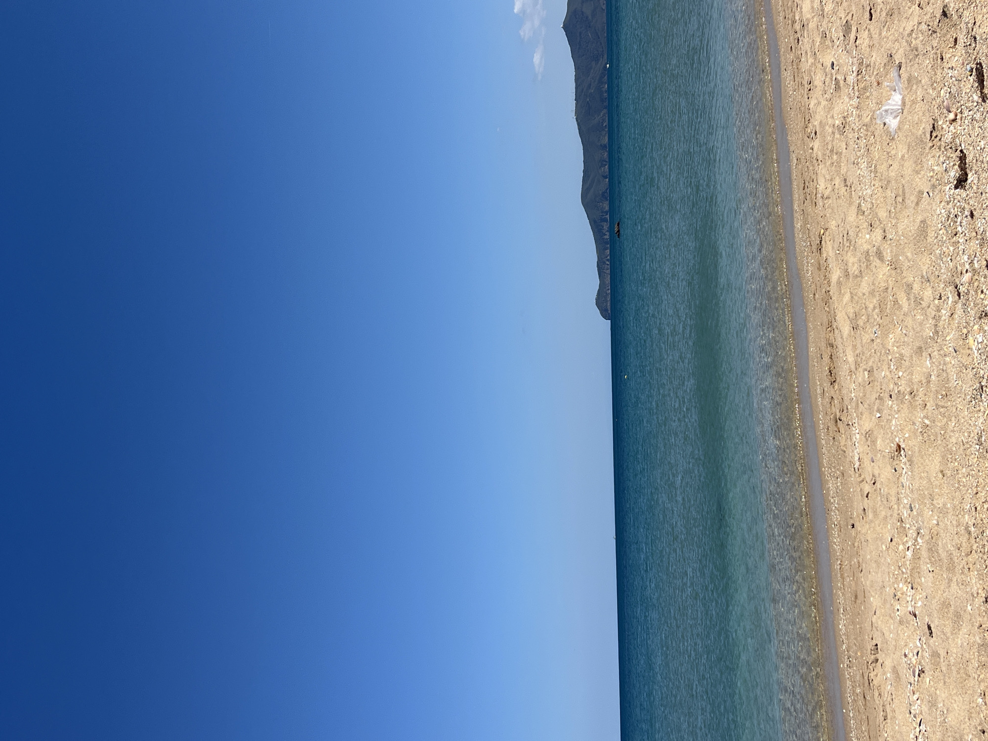 The image depicts a coastal scene in M'Diq, Morocco. The scene shows the beach with sand and some shells, extending into the sea. In the background, there's a mountain range visible. A plastic bag can be seen on the beach. The sky is clear with blue.