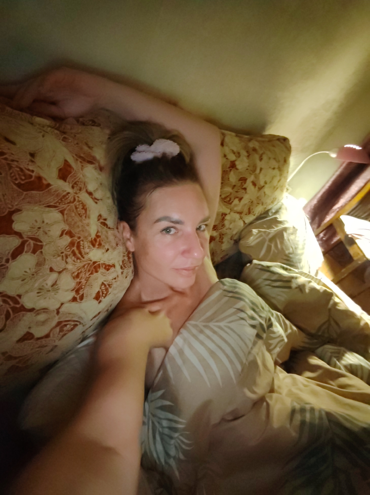 A woman is lying in bed under covers, presumably indoors. She is propped up by pillows. Her hair is pulled back with a hair accessory. A hand is resting on her chest or shoulder. The bed has patterned bedding and pillows. The location is not discernible from the image.