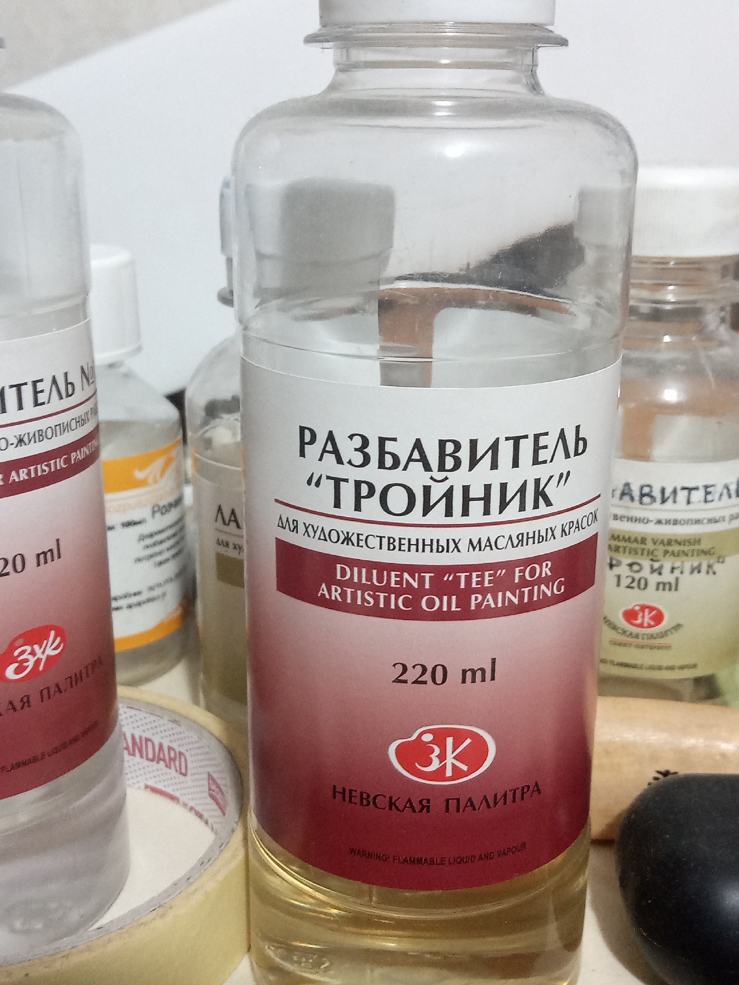 The image shows a close-up of several bottles of art supplies, primarily oil painting mediums and solvents, arranged on a white surface. The dominant bottle in the foreground is a clear glass bottle with a white cap, containing a pale yellow liquid. It has a label with red and pink accents. The label text is in Russian and English. The Russian text at the top reads "РАЗБАВИТЕЛЬ" and "ТРОЙНИК". Below this, in English, it says "DILUENT 'TEE' FOR ARTISTIC OIL PAINTING". Further down, it indicates "220 ml" and displays a red oval logo with "ЗК" inside. At the very bottom of the label, smaller text reads "WARNING! FLAMMABLE LIQUID AND VAPOUR".
To the left of the main bottle is another similar clear bottle, also partially visible, with a label that is mostly obscured but shows "20 ml" and a similar red oval logo. Behind the main bottle, to the right, is a smaller bottle with a clear liquid and a label that is too blurry to read clearly, though some text appears to be in English and Russian. To the far right, out of focus, is a cylindrical object, possibly a wooden brush handle or a part of another art tool, and a dark, round object, perhaps a piece of charcoal or a stopper. There is also a roll of beige masking tape with red and white markings visible in the lower left corner of the image. The background is a plain white surface, likely a table or shelf. There are no people or animals present in the image. The lighting is bright, suggesting an indoor setting.