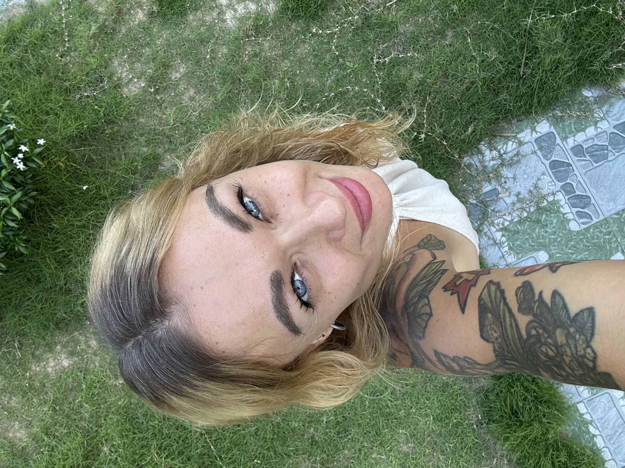 A high-angle selfie captures a woman looking upwards directly at the camera, with a slight smile. Her face is the central focus, framed by her blonde hair with visible darker roots. She has striking blue eyes, dark eyebrows, and wears pink lipstick. Small, silver hoop earrings are visible on her ears.
A prominent, colorful tattoo covers her right arm and shoulder, depicting intricate green and darker patterns resembling foliage or organic designs, with hints of red and yellow. She is wearing a light-colored, possibly white or cream, top.
The background primarily consists of green grass, some patches appearing drier or sparser. In the top left, there's lush green foliage adorned with small white flowers. The bottom right of the image reveals a section of a light gray-green tiled pathway or patio, featuring an interlocking geometric pattern. The scene is brightly lit by natural daylight, suggesting an outdoor setting during the day, consistent with the provided location context of Mũi Né, Vietnam. No text is visible in the image.