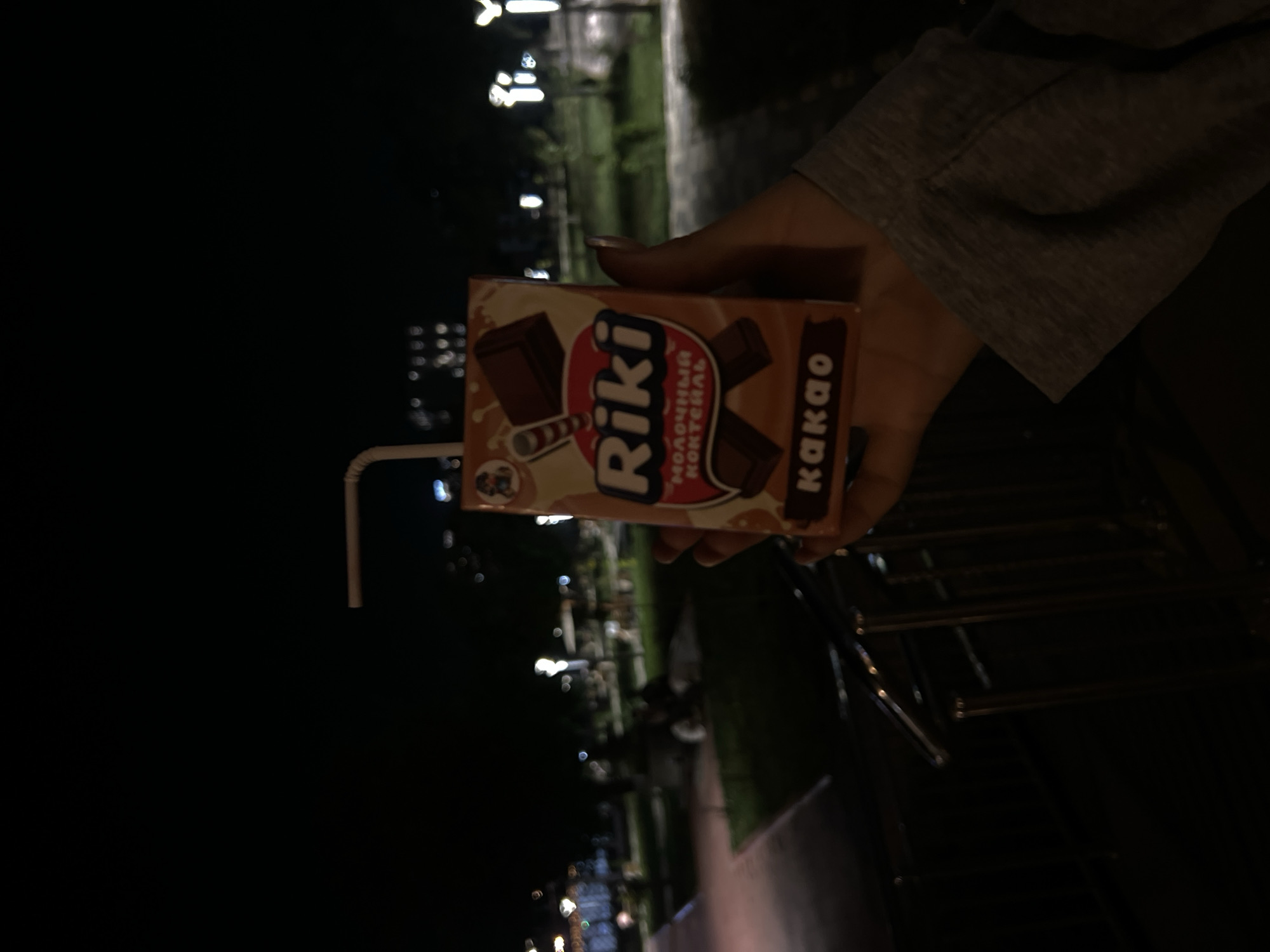 A person's hand, wearing a grey long-sleeved shirt, holds a chocolate milk juice box labeled "Riki Kakao" in front of a dark, outdoor environment. The background consists of blurred park lights and foliage, suggesting a nighttime setting. The scene is shot from a slightly low angle.