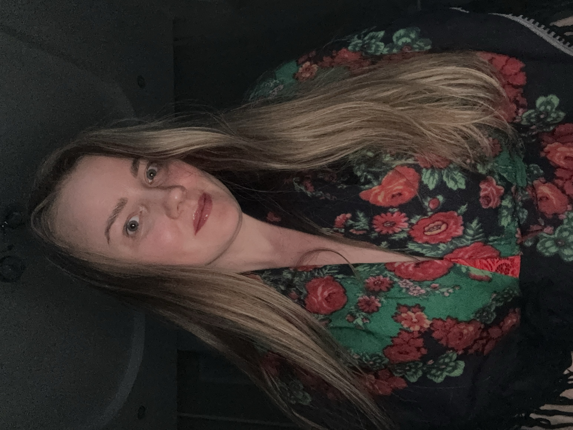 A fair-skinned female with blonde hair is centered in the image, wearing a floral-patterned garment with roses and green leaves on a dark background. A black shawl with fringes is partially visible on the right side. The background appears to be a dark, possibly interior space. There is no explicit indication of Chaikovsky, Russia in the image.