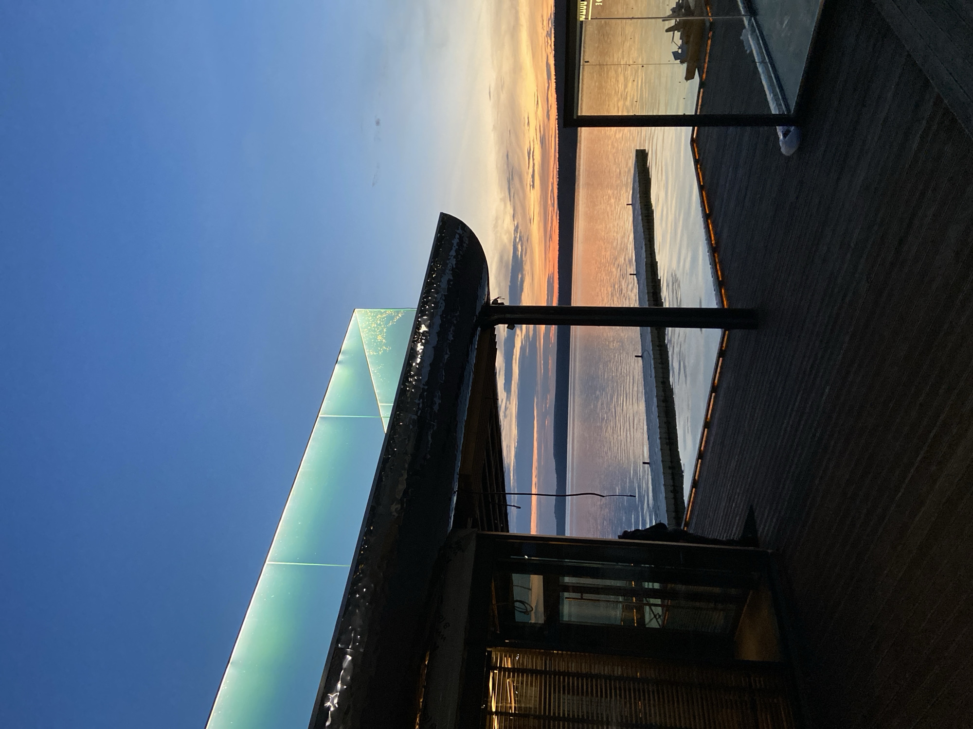 The image depicts a waterfront scene at sunset, likely in Votkinsk, Russia. The sky is a gradient of deep blue fading into warm oranges and pinks, indicative of the golden hour. The sun is setting over a body of water, casting a warm glow and reflections.
In the foreground, a modern structure with a sleek, angled glass facade is visible. This building features dark, angular framing, and the glass appears to have a slight greenish tint, reflecting the sky and surrounding elements. Adjacent to this is a dark wooden deck or walkway, with visible planks and subtle accent lighting along the edge facing the water.
Extending into the water from the deck are dark posts supporting what appears to be a pier or walkway. In the distance, across the water, a tree line is discernible against the colorful sky. The water itself is calm, with subtle ripples and reflections of the sky and structures. There are also some distant boats or structures visible on the water in the far distance, adding to the sense of a developed waterfront area.
The overall mood is serene and tranquil, enhanced by the beautiful sunset and the peaceful water. The architecture suggests a contemporary design, possibly a restaurant, cafe, or public space designed to take advantage of the waterfront views. No people are clearly visible in the image. The scene is captured from a slightly elevated perspective, looking out over the water.