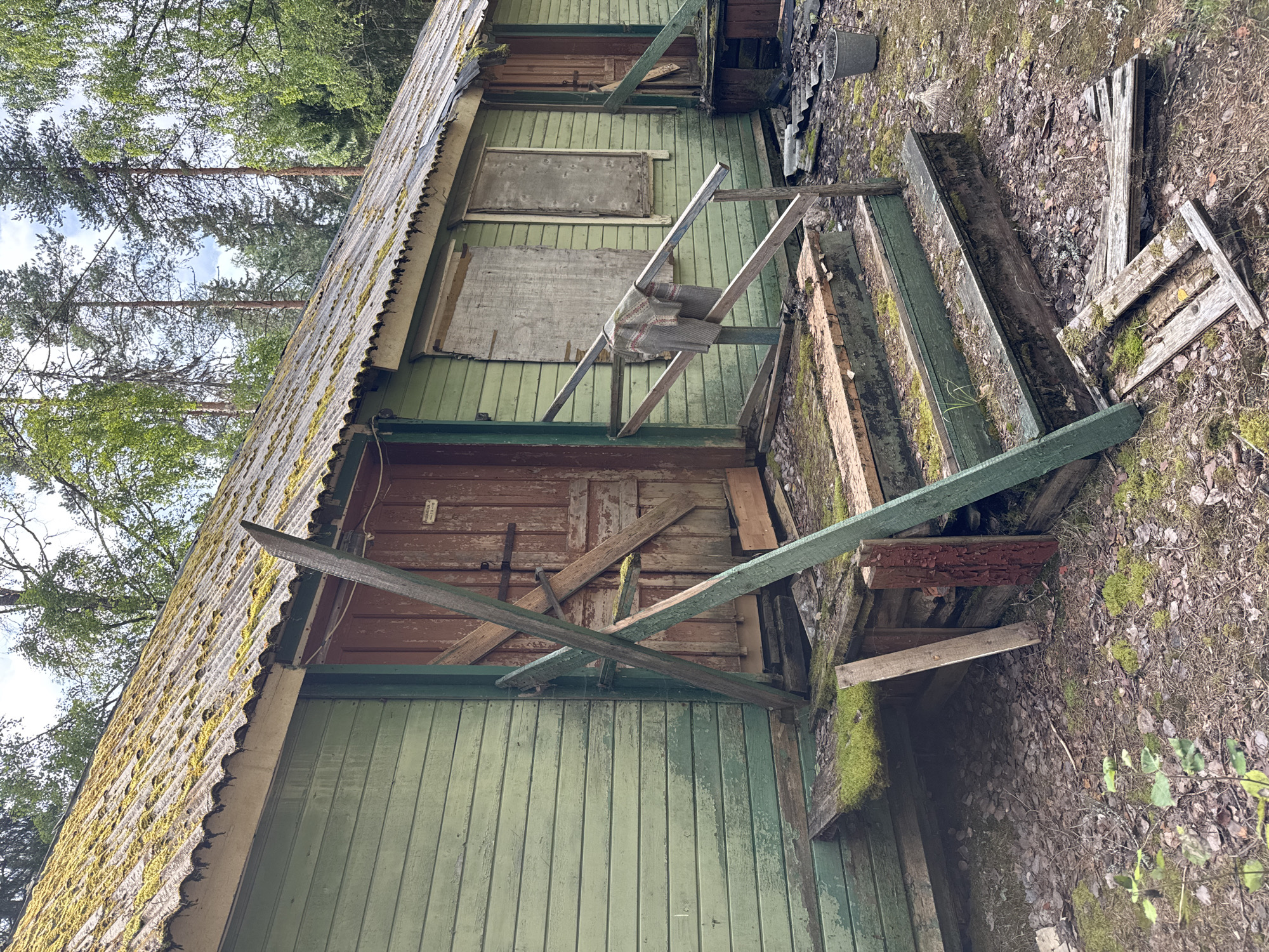 The image depicts a dilapidated, abandoned wooden building with light green siding and a corrugated metal roof covered in moss. The building appears to be a long, single-story structure with a weathered wooden door in the center and boarded-up windows. The porch area is in disrepair, with missing and broken wooden planks. There is a wooden railing on the right side of the porch, partially covered by a grey and red knitted sweater. Scattered debris, including broken wood and what appears to be a metal bucket, litters the ground around the building. The environment is overgrown with vegetation, with trees visible in the background. The scene suggests a rural or semi-rural setting, likely in a wooded area. The location is identified as Ikhala, Russia.