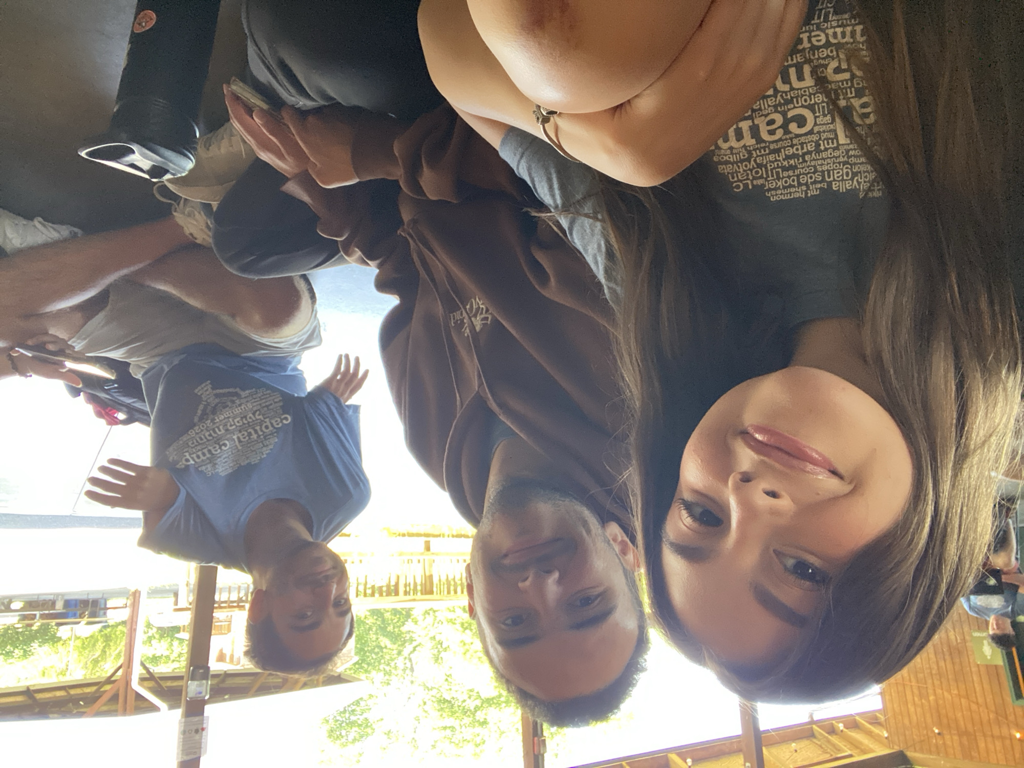 Three people are seated on the ground in front of a wooden structure with a covered roof, suggesting an outdoor recreational area. The person on the left, a young woman, is looking at the camera with a smile. She has long brown hair and is wearing a gray t-shirt with text and graphics. The person in the middle, a young man, is looking slightly away from the camera and is wearing a brown hooded sweatshirt. The person on the right, a young man, is looking towards the camera and gesturing with his hands. He is wearing a blue t-shirt with text and graphics and white shorts. A black water bottle is visible in the foreground. The background shows more wooden structures, trees, and a bright sky. The overall scene appears to be a casual gathering in an outdoor setting.