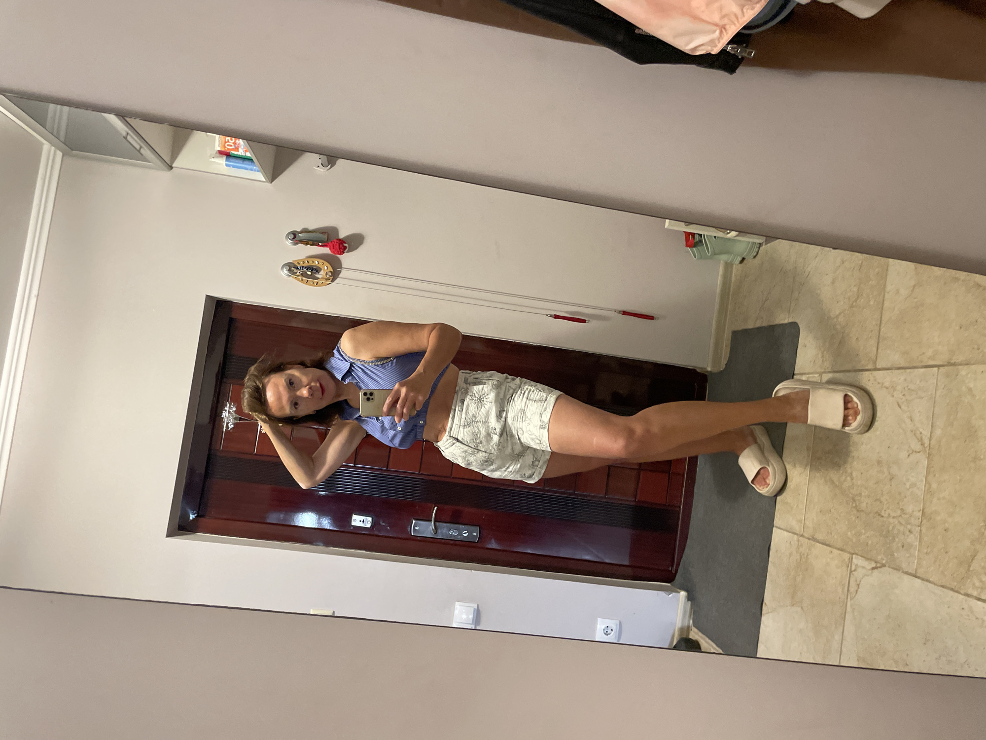 A woman is standing in what appears to be a hallway and taking a mirror selfie. She is wearing a blue cropped button-up shirt and white shorts with a subtle pattern. She has her hair up and is wearing white slide sandals. In the background, a dark brown wooden door with a silver handle is visible. The hallway has beige tiled flooring and a grey mat in front of the door. On the wall to the right of the door, there are a couple of decorative items, one with red tassels. Further to the right, a tall mirror reflects the scene, and a shelf above the mirror holds a few items including what looks like bottled drinks and books. A pink jacket is visible hanging on the right side of the mirror frame, and the reflection shows a sliver of a door and the wall beyond. The environment suggests a residential interior, possibly an apartment or house hallway.