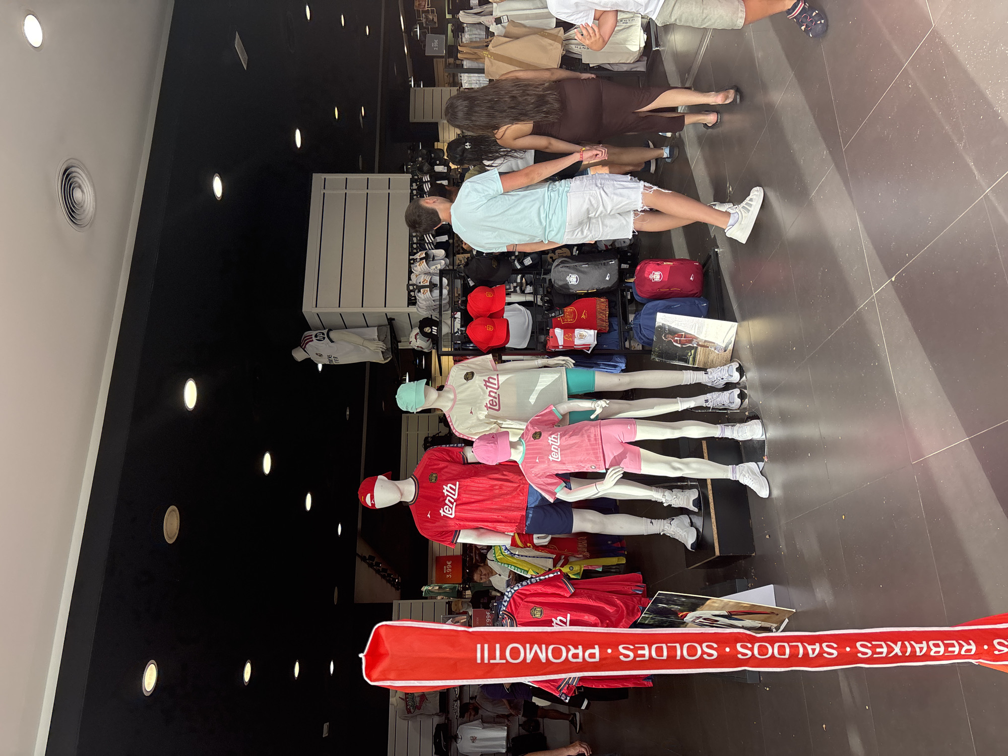 A storefront in Orihuela, Spain, displays athletic apparel on mannequins. The mannequins, a tall adult male, a child, and two adult females, are dressed in t-shirts, shorts, and hats, predominantly in red and pink. Several red soccer jerseys are also displayed. A large red banner with white text reads "S. REBAIXES • SALDOS • PROMOTII". In the background, shelves are stocked with hats, bags, and other merchandise. Three young people, two males and one female, are walking away from the display, one male holding hands with the female. The flooring is tiled, and the lighting appears to be from recessed ceiling fixtures.
