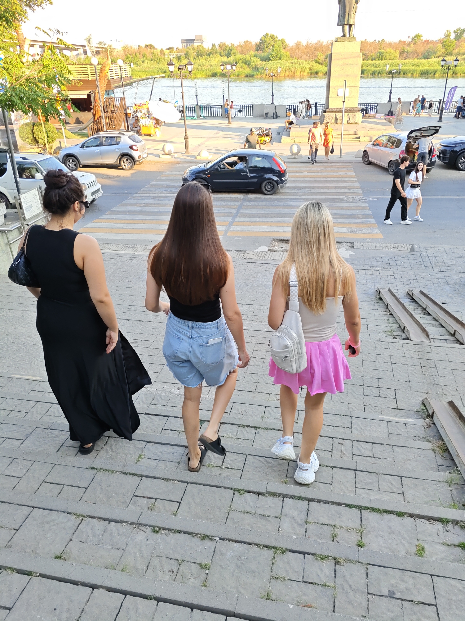 Three women are walking down a set of stone stairs in Rostov-on-Don, Russia. The woman on the left is wearing a long black dress and carrying a black handbag. The woman in the middle is wearing a black tank top and denim shorts. The woman on the right is wearing a light-colored tank top and a pink skirt, carrying a light-colored crossbody bag. In the background, a body of water, possibly the Don River, is visible, with trees and buildings lining the opposite bank. Several cars and pedestrians are also present in the background, indicating a public area. The overall scene suggests a warm day in a city park or waterfront promenade.