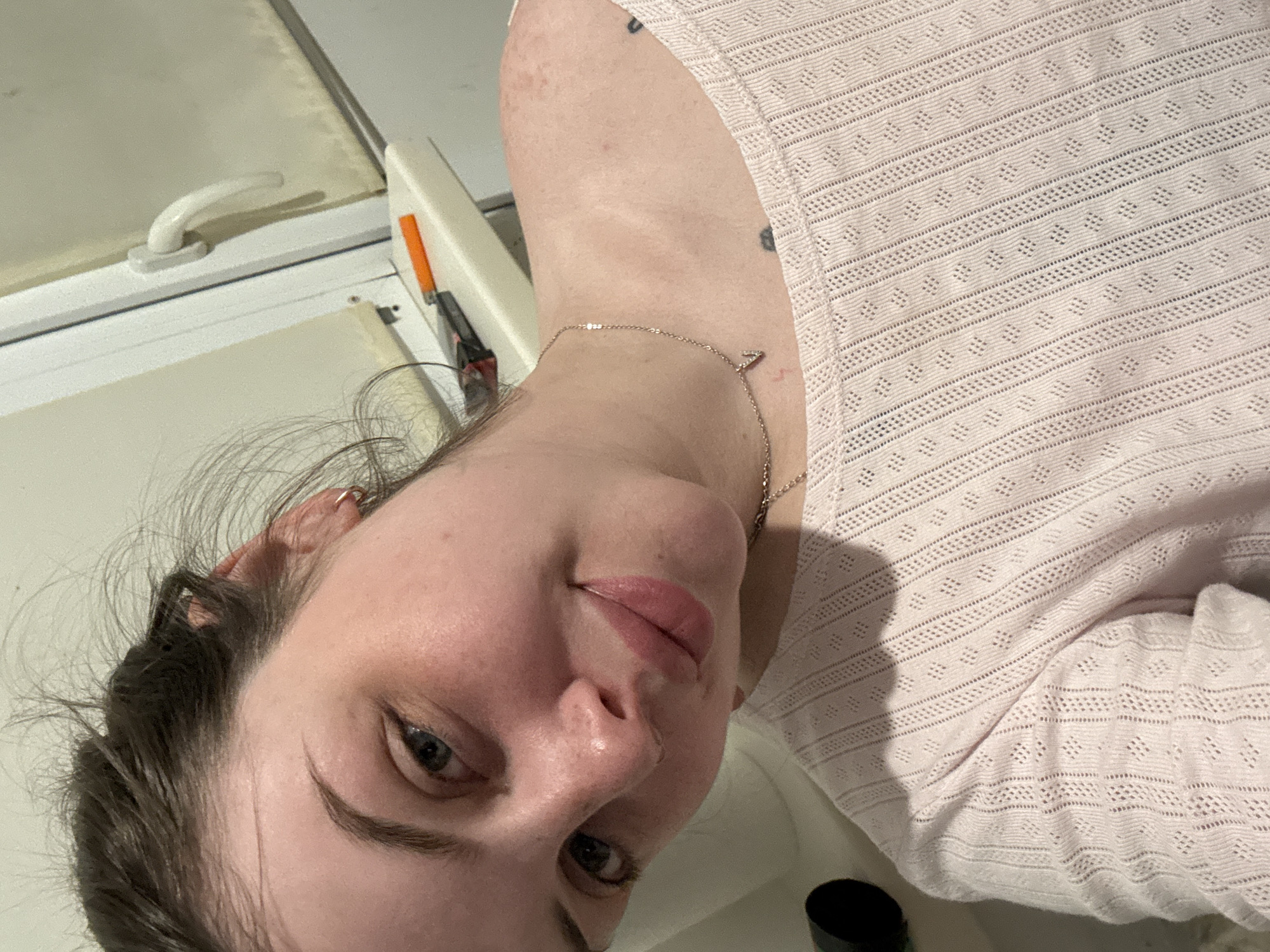 A selfie taken indoors features a fair-skinned person with dark hair pulled back, wearing a light pink open-weave shirt and a delicate gold necklace with a "V" pendant. The person is angled towards the camera with their head tilted. The background includes a white window frame with a handle, suggesting an interior space. The location in Chornomorskoe, Ukraine is not evident from the image.