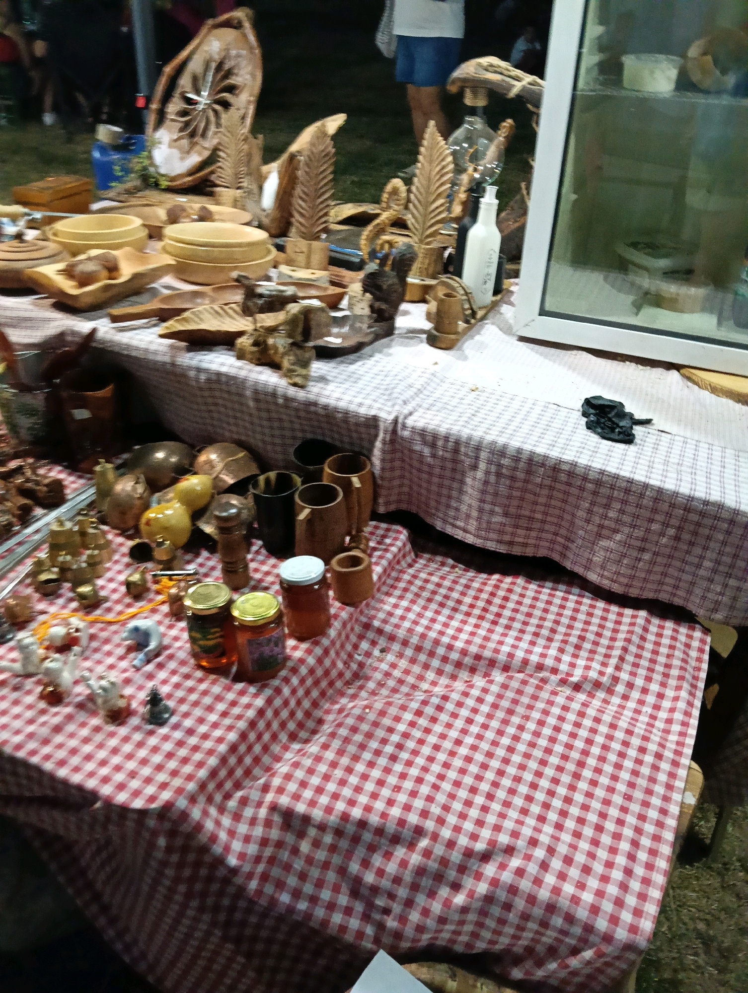 The image depicts a market stall with various handcrafted wooden items and jars of preserves displayed on tables covered with red and white checkered tablecloths. In the background, a glass display case is visible, containing more items. The overall setting appears to be outdoors at night, suggested by the lighting and dark background. There are no people clearly visible in the immediate foreground, but a few figures can be vaguely discerned in the background, indicating a public gathering or event. The items on display include carved wooden bowls, decorative wooden sculptures resembling leaves and feathers, and small decorative figurines. There are also several glass jars filled with amber-colored preserves, likely honey or jam, topped with metal lids. The scene is arranged in a way that suggests a rustic or artisanal market.