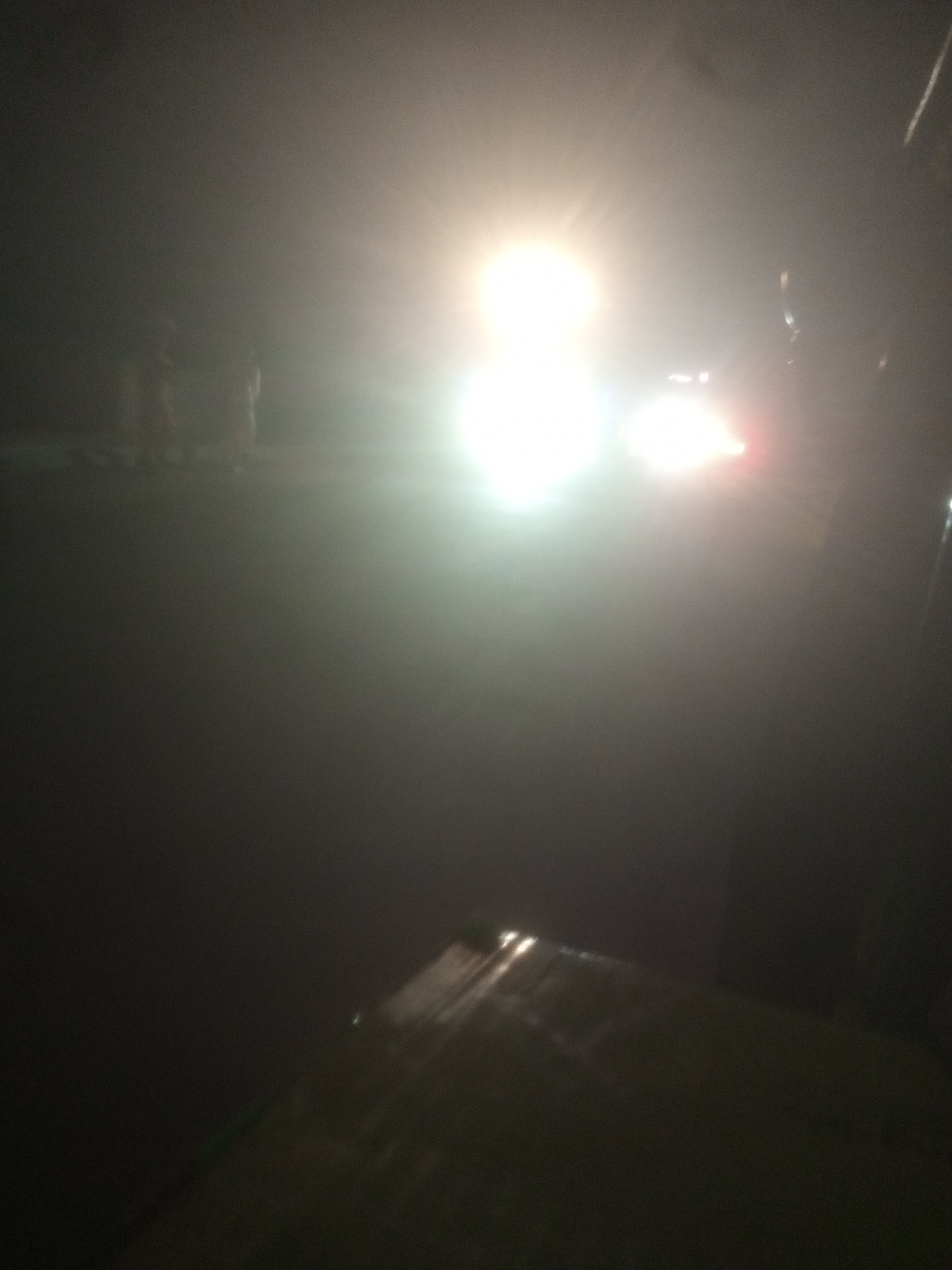The image depicts a foggy or hazy nighttime scene, likely on a road, with prominent vehicle headlights. The lighting suggests a low visibility condition. In the background, faint silhouettes of several individuals are visible, appearing to be standing or walking. The headlights of what appears to be a car or a set of vehicles are a bright, white glare in the center of the frame, with red taillights visible to the right of the main glare. The foreground is dark and out of focus, with a diagonal object that may be a surface or structure. The image is grainy, indicating it was likely taken with a mobile phone camera in low light conditions. The location is identified as Tundun Wada West, Nigeria, suggesting an urban or semi-urban road environment.
