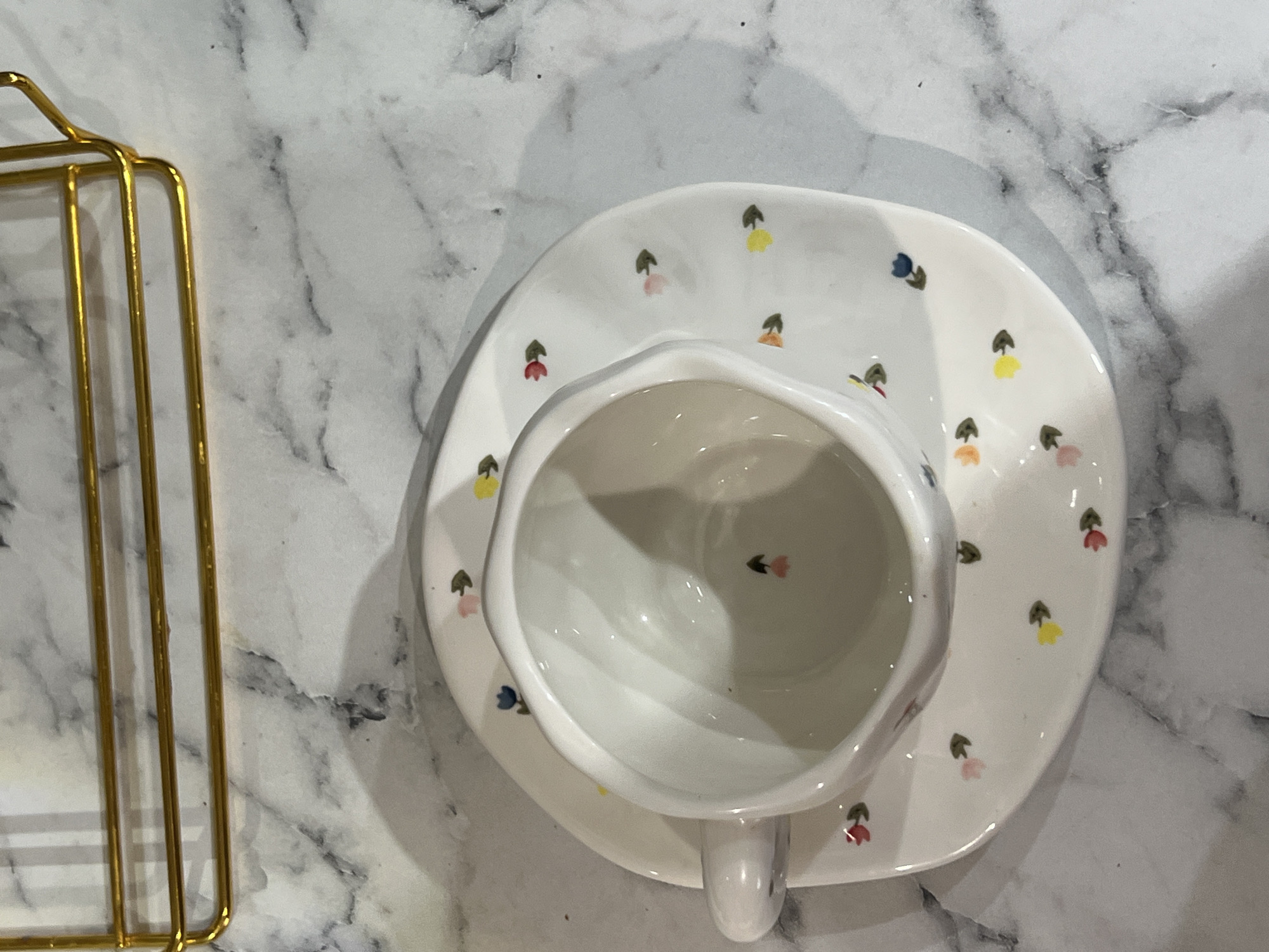 A top-down shot shows a white teacup and saucer with a floral pattern on a marble-like surface. To the left, part of a gold-colored metal stand is visible. The teacup is empty and centrally positioned on the saucer.