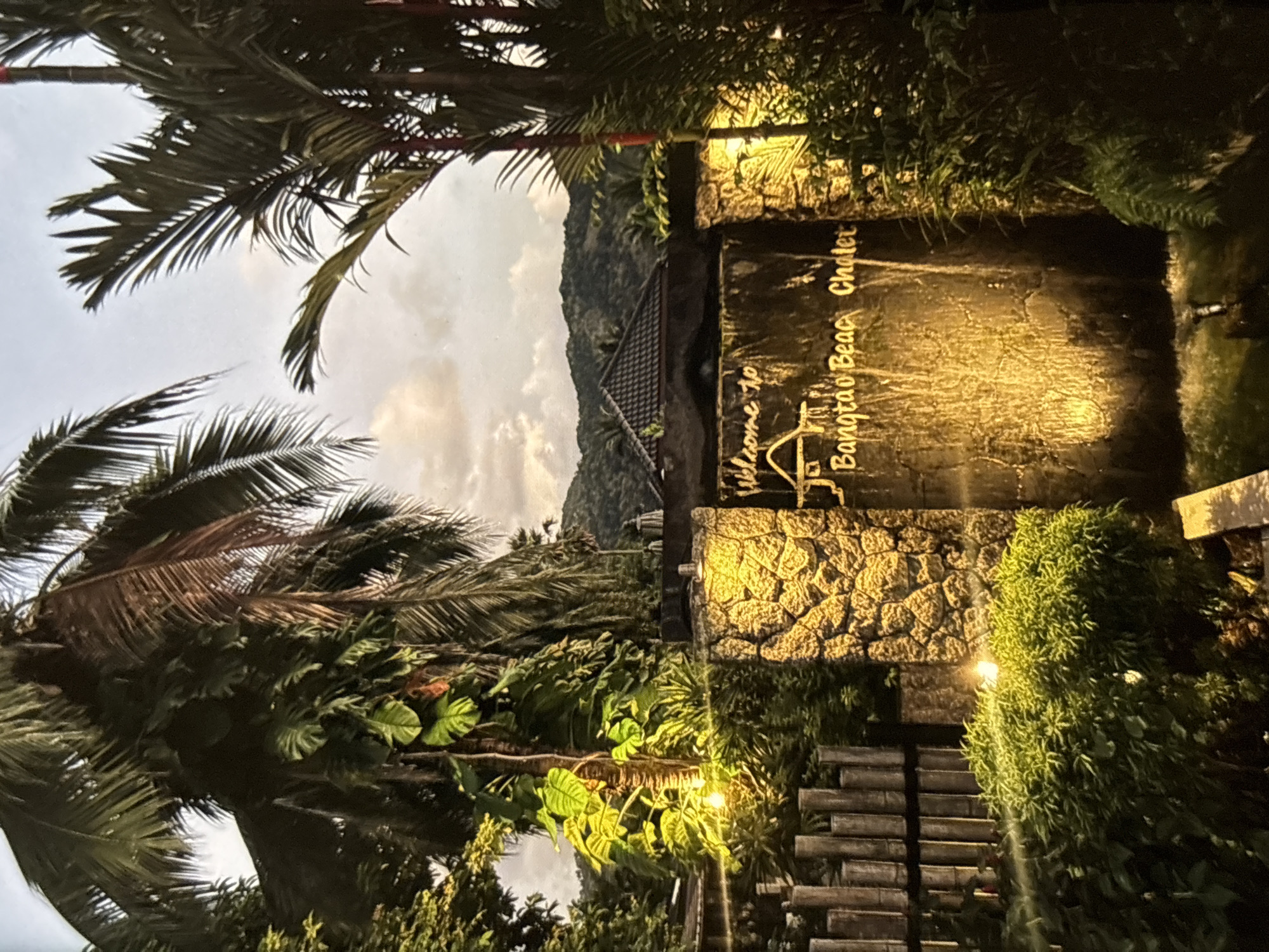 The image depicts a tropical entrance at dusk. A stone wall with a cascading waterfall feature is illuminated by warm, yellow lights, illuminating the text "Welcome to Bangtao Beach Chalet" in gold lettering. The wall is flanked by lush green foliage and palm trees. In the background, a dark, mountainous landscape is visible against a cloudy sky. To the left, a bamboo fence and more greenery are illuminated by smaller lights. No people are visible in the scene.