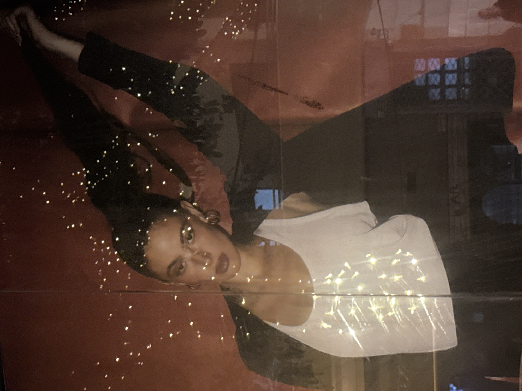 A medium shot depicts a woman lying on a reddish-brown surface. She has dark hair and fair skin. She is wearing a white tank top and a black jacket or top. Her arms are extended, one reaching towards the top left corner of the frame and the other towards the upper right corner. The background reflects a dimly lit interior with what appears to be a grid pattern resembling a window or glass facade. There are scattered bokeh lights throughout the scene, adding a soft, diffused lighting effect. The location appears to be a photograph or print of someone in Ramat Gan, Israel.