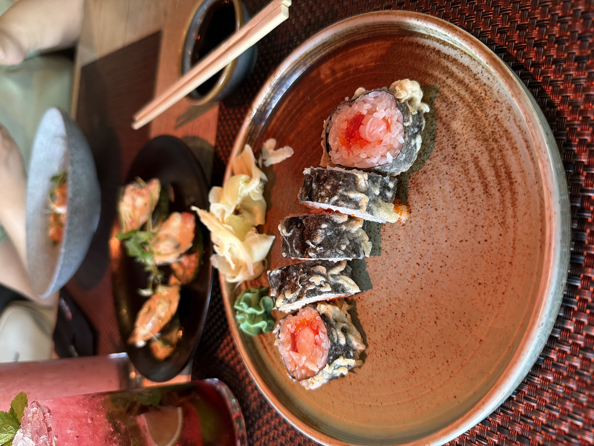 A close-up shot captures a plate of deep-fried sushi rolls, garnished with a small mound of bright green wasabi and pickled ginger, set against a textured brown placemat. In the background, a black plate holds what appears to be a serving of mussels, and further back, a grey bowl contains a colorful dish, possibly a salad or seafood appetizer. A tall glass filled with a pink beverage, ice, mint, and lime wedges sits to the left. Wooden chopsticks rest above a small bowl of dark liquid, likely soy sauce, in the upper right quadrant of the image. The scene suggests a dining setting, possibly in a restaurant in Rostov-on-Don, Russia, given the details and the overall presentation.