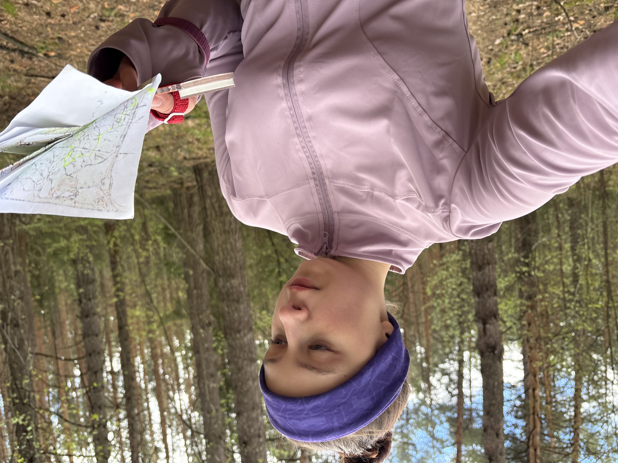 A young woman is standing in a forest. She is wearing a lavender zip-up jacket and a purple headband. She is holding a map and a compass. The trees are tall and thin. The location is likely in the city of Perm, Russia.