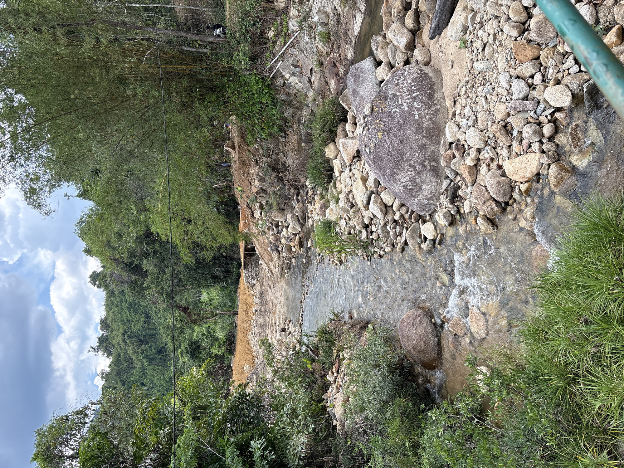 The image shows a stream running through a rocky area with lush vegetation. The location is Khanh Vinh, Vietnam. The stream bed is filled with rocks and boulders, and the water is clear and shallow. Green vegetation lines the banks of the stream, including trees, bushes, and grass. There is evidence of some construction or excavation along the stream bank. In the background, there are dense trees, and a partly cloudy sky is visible. Two motorbikes are parked on the bank far in the background.