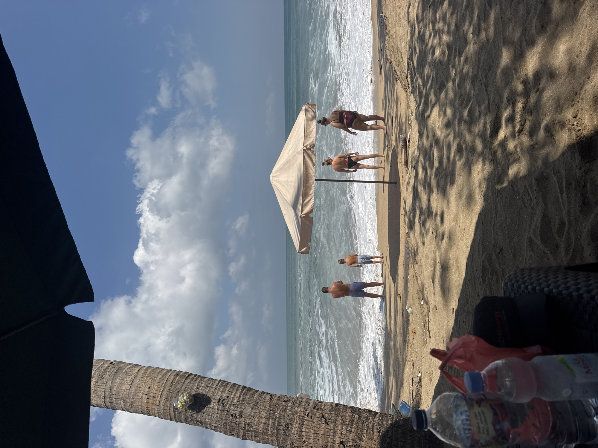 A family stands near the ocean on a sandy beach in Kuta, Indonesia. In the foreground, a palm tree trunk is on the left. In the middle ground, a man and a boy stand facing the ocean. To their right, two women stand under a beige umbrella, facing the ocean. The ocean is rough with waves. In the foreground, there are two water bottles and a red plastic bag on a textured surface. The sky is blue with scattered white clouds.