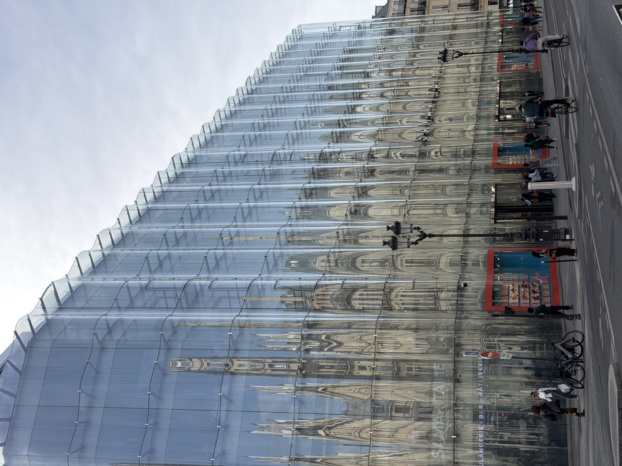 The image displays a modern, multi-story building with a distinctive wavy, reflective glass facade, towering over a bustling urban street. The glass reflects a traditional, ornate building (likely Haussmannian architecture) across the street, creating a distorted, dreamlike mirror image of its intricate details, along with the bright, overcast sky.
At street level, a paved road runs parallel to a sidewalk. Several people are visible, engaged in various activities: some are walking along the sidewalk, while others are on bicycles or scooters on the street. Two bicycles are parked on the sidewalk. Street infrastructure includes a black lamppost, a white information pillar, and a red circular traffic sign. The building's lower section features red rectangular window displays that contrast with the reflective facade.
The scene depicts a daytime setting, characterized by bright but diffused light, suggesting an overcast sky. The visible reflections and architectural styles strongly indicate a European city. Text reflected on the building's facade clearly spells out "SAMARITAINE" and "BOULANGERIE • É • BAR • PÂTISSERIE", identifying the building as La Samaritaine department store in Paris, France.