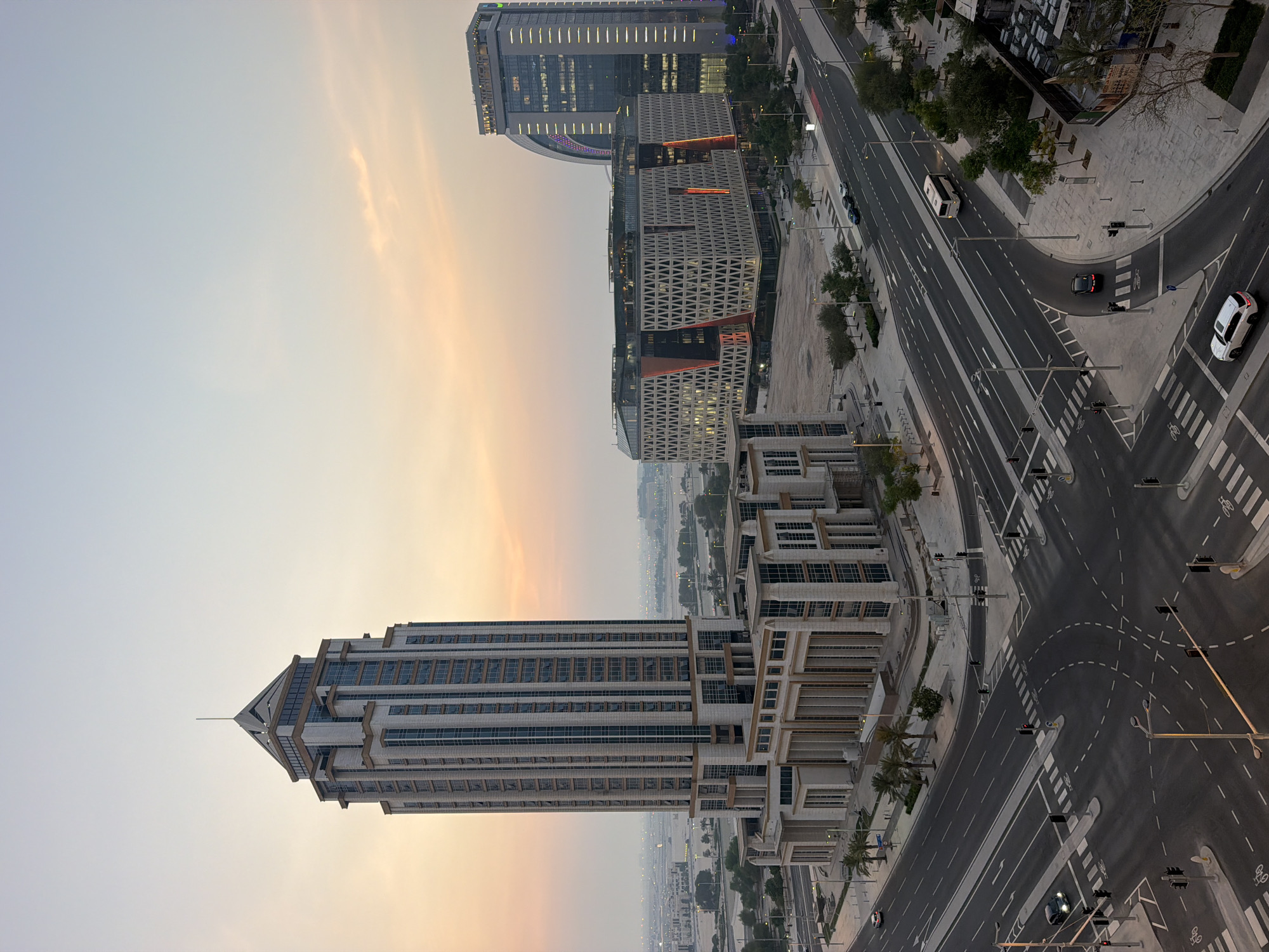 This is a high-angle, full shot of a modern city intersection in Lusail, Qatar, during the golden hour. The sky is a soft gradient of pale blue and orange hues, indicating either sunrise or sunset. In the foreground, a complex, multi-lane intersection is visible, marked with white dashed lines and pedestrian crossings. Traffic lights are positioned at various points around the intersection.
Dominating the left side of the frame is a tall, multi-story skyscraper with a distinct pyramidal roof. Its facade is characterized by a grid of windows and beige vertical and horizontal accents. The lower section of this building appears to be a more complex structure with colonnades.
To the right, two other contemporary buildings are prominent. One is a lower, wider structure with a distinctive geometric facade featuring a pattern of interlocking triangles and illuminated red vertical accents. Adjacent to it, a taller, slender skyscraper rises, its facade featuring vertical lines of illuminated lights and a curved upper section.
Several vehicles are present on the roads: a white bus is moving along one of the lanes, a black car is seen turning, and a white SUV is parked near the intersection. A few cars are also visible further in the distance. Palm trees and other greenery are interspersed throughout the scene, lining the roads and sidewalks. In the background, a hazy cityscape stretches towards the horizon, with other buildings and structures faintly visible. The overall impression is one of a well-developed, modern urban environment.