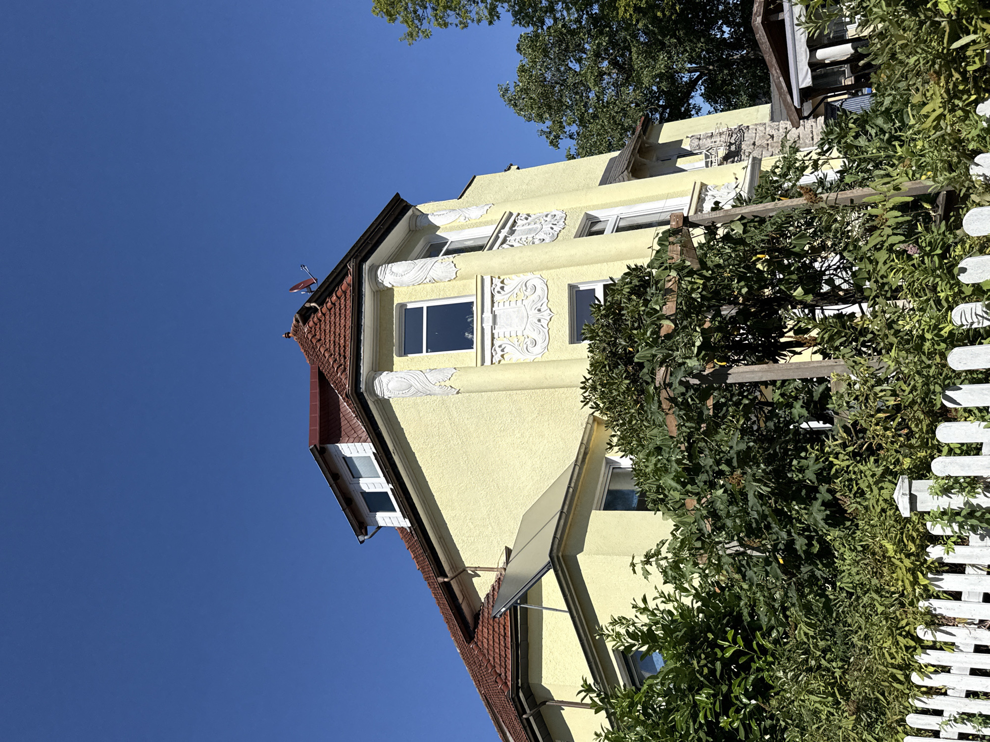 This is a medium-shot, eye-level, outdoor, daytime, full-color photograph of a light yellow, multi-story building with a red tile roof, set against a clear blue sky. The building features decorative white stucco elements, including columns and ornate designs above windows. A solar panel is visible on the roof, and a small satellite dish is mounted on the peak of the roof. The foreground is partially obscured by dense green foliage, including trees and shrubs, and a white picket fence runs along the bottom of the image. A wooden pergola structure is also present, entwined with vegetation. The architecture suggests a European style, potentially from the early to mid-20th century. Based on the context provided, this building is located in Bad Sachsa, Germany.