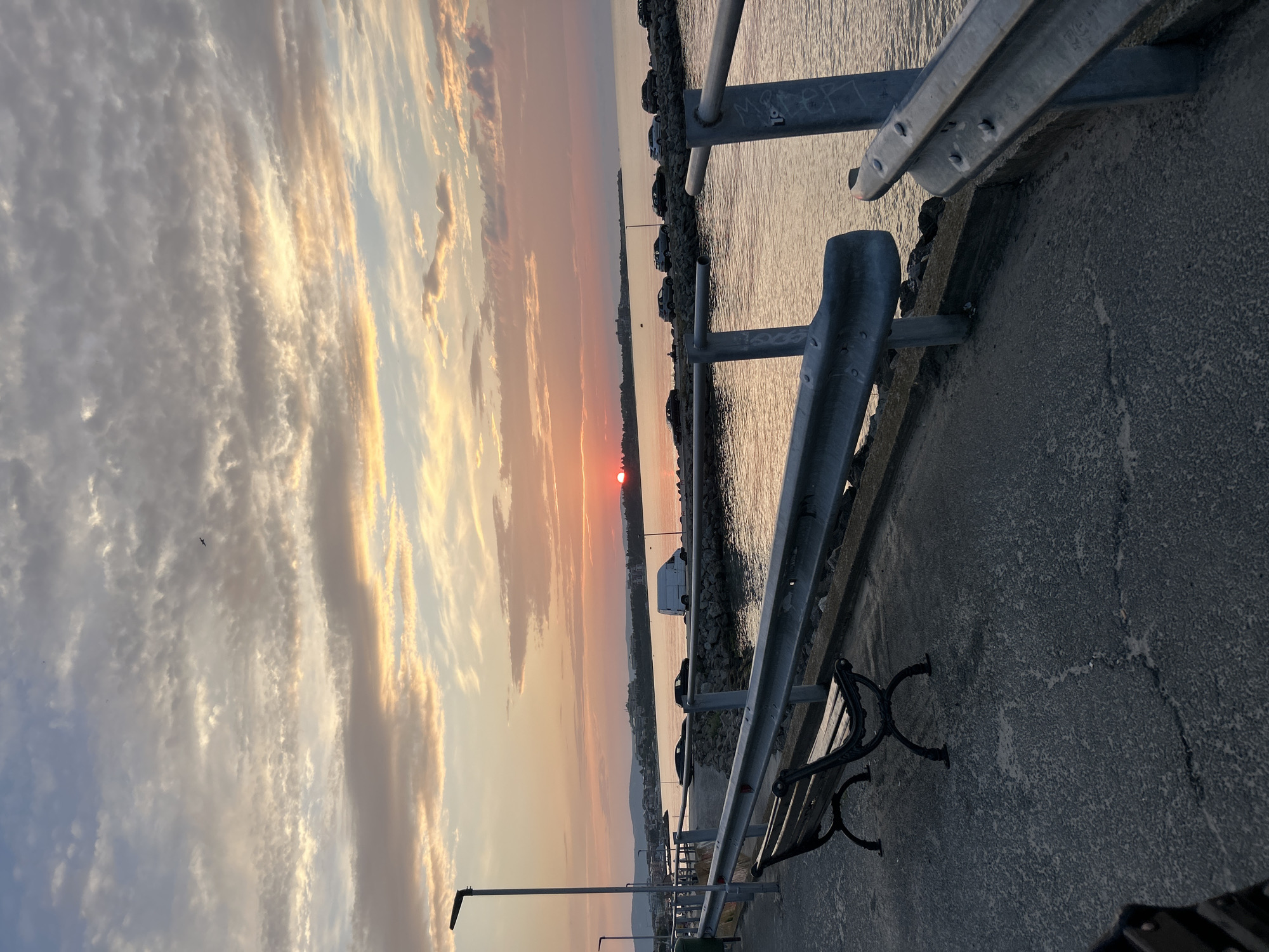 The image depicts a serene sunset over the Black Sea in Ravda, Bulgaria. A paved walkway runs along the left side of the frame, featuring a weathered park bench. A metal railing separates the walkway from the water's edge, where a breakwater made of rocks is visible. Several cars are parked along the breakwater, and a white van is also present. The sky is painted with hues of orange, pink, and yellow as the sun dips below the horizon, casting a warm glow over the tranquil sea. The distant coastline is lined with buildings, suggesting a coastal town. The overall atmosphere is peaceful and picturesque, capturing the natural beauty of a sunset by the sea.