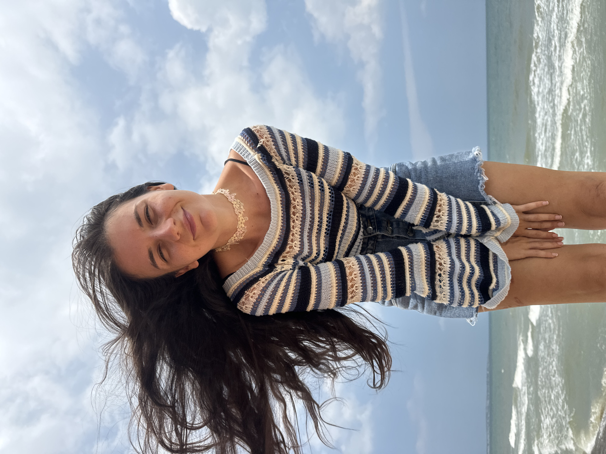 A young woman with long, dark hair is standing on a beach in Mũi Né, Vietnam, with the ocean and a cloudy sky in the background. Her hair is blowing in the wind, framing her face as she smiles with her eyes closed. She is wearing a striped, crochet sweater with blue, white, and tan stripes, and a denim skirt. A delicate, cream-colored choker is around her neck. The waves of the ocean are visible in the lower part of the frame, with the shoreline in the foreground. The overall scene conveys a sense of joy and freedom.