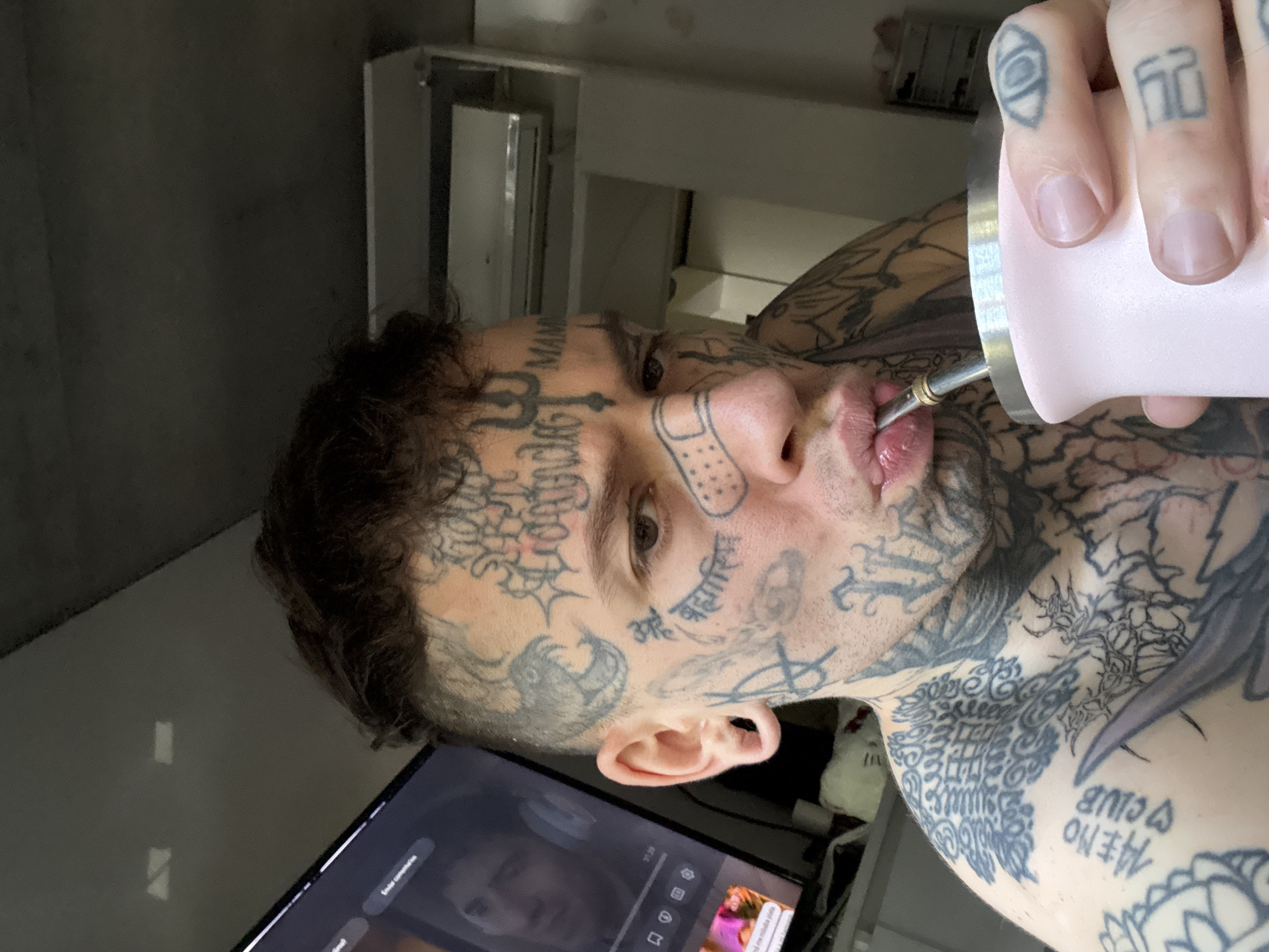 An adult male, heavily tattooed across his face, neck, shoulders, and upper torso, is depicted in an indoor setting. His short, dark hair is styled with shaved sides, and he has stretched earlobes. Notable facial tattoos include a band-aid graphic on his nose bridge, a trident symbol on his forehead, "MAMBA" on his right temple, and various intricate symbols and text. His left index finger also bears an eye tattoo, and "MEMO CLUB" is tattooed on his left shoulder. He holds a light pink mate cup with a silver rim in his left hand and is shown with his lips pursed around a metallic bombilla, indicating he is drinking from it. He is looking directly at the camera. In the background, a television screen on the left displays a video featuring a man's face and the text "Enviar comentarios" (Send comments), with the time "17:29" visible. To the upper right, a white wall-mounted air conditioning unit is present. The environment consists of plain, light-colored walls and a ceiling, suggesting a private residence. The cultural elements, such as the mate cup and Spanish language on the television, are consistent with a dwelling in Buenos Aires, Argentina.