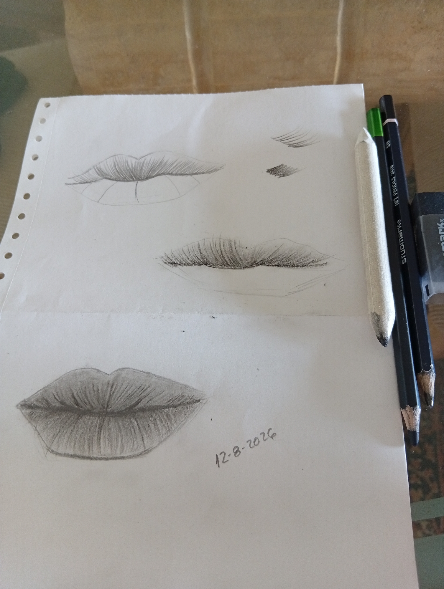A pencil sketch of lips and eyelashes is displayed on a sheet of white paper. The paper is positioned on a wooden surface. Three pencils and an eraser are placed to the right of the paper. The sketch includes two smaller drawings of lips and eyelashes in the upper section of the paper, and a larger, more detailed drawing of a mouth in the lower section. A date, "12-8-2026," is written on the paper near the bottom right corner. The overall scene is an indoor setting, likely a home or studio, given the wooden table and the casual arrangement of art supplies. There is no indication of San Antonio de Los Altos, Venezuela, in the image.