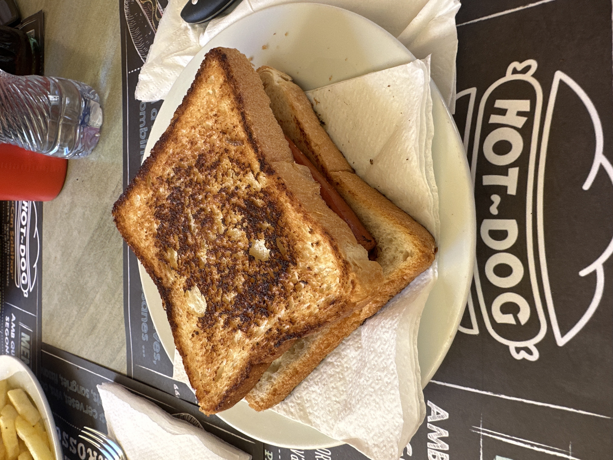 A close-up, eye-level shot shows a toasted sandwich with a hot dog inside, served on a white plate with a white napkin. The sandwich is the main focus, with its golden-brown, crispy toast and visible filling. The plate rests on a black placemat featuring a white "HOT DOG" logo. In the background, a white napkin and a fork are visible on the left, while a red cup, a plastic water bottle, and a dark beverage are on the right. The setting appears to be an indoor eatery, likely a cafe or restaurant, in Sant Feliu De Guíxols, Spain, suggested by the Spanish text on the placemat.